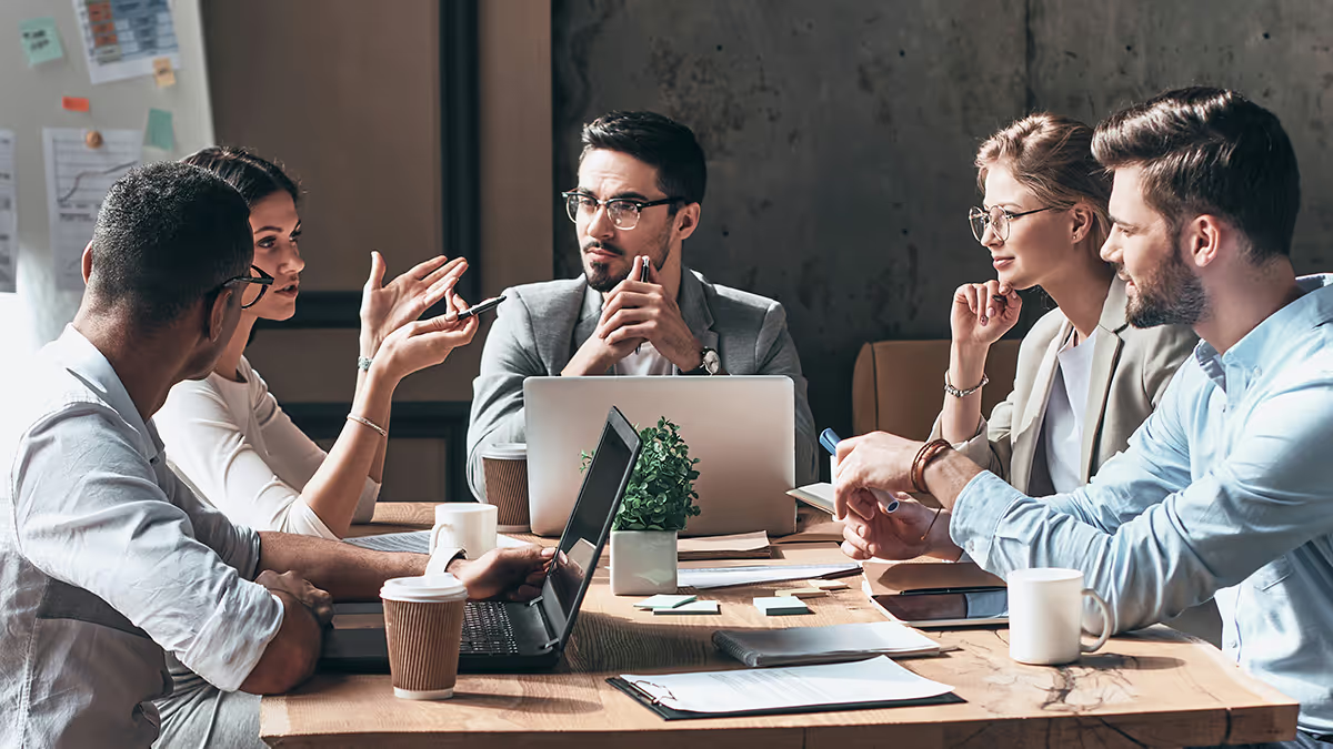 Five professionals in animated discussion around a wooden conference table with laptops