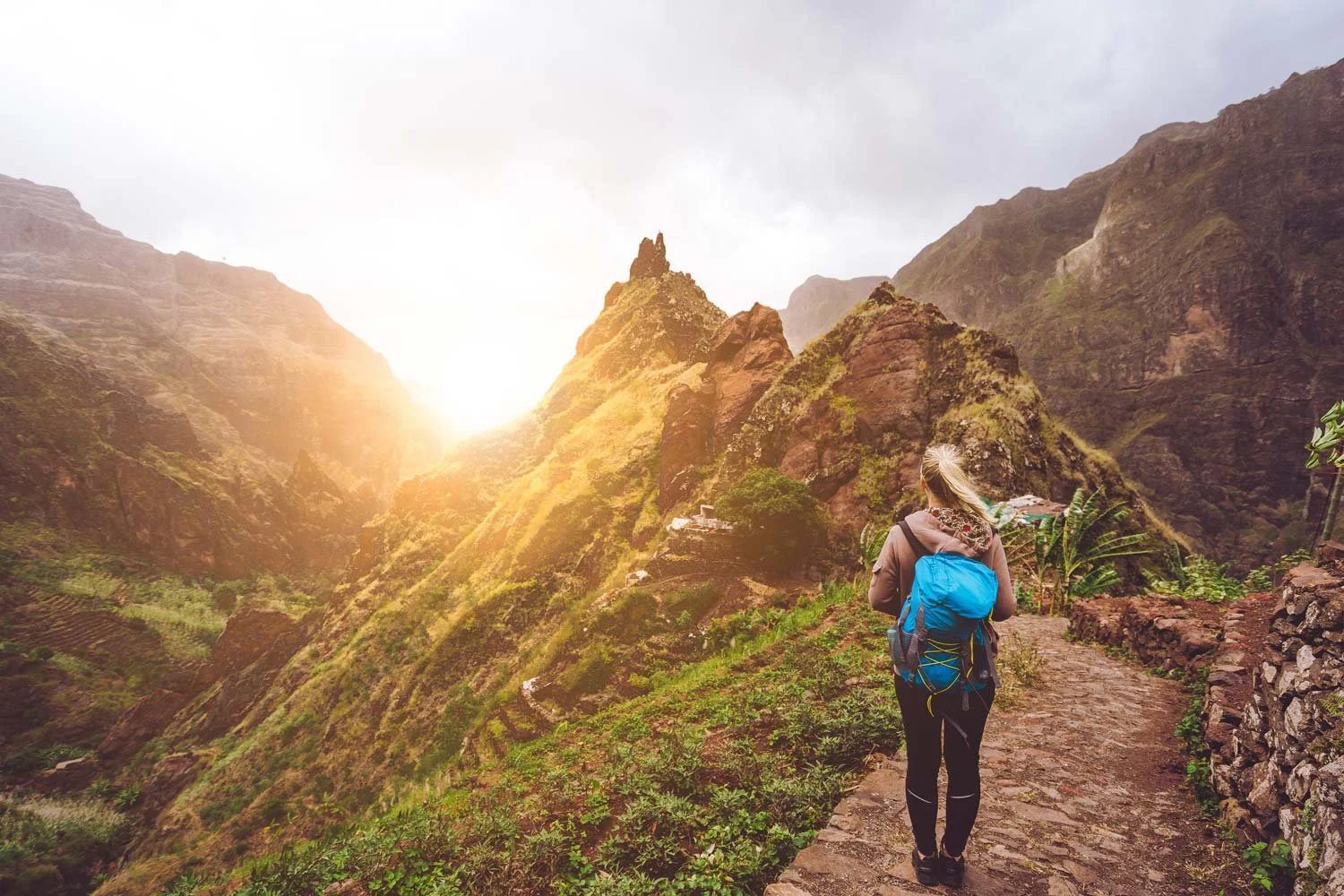Solo female hiker with a blue backpack walking a stone trail toward sunlit peaks