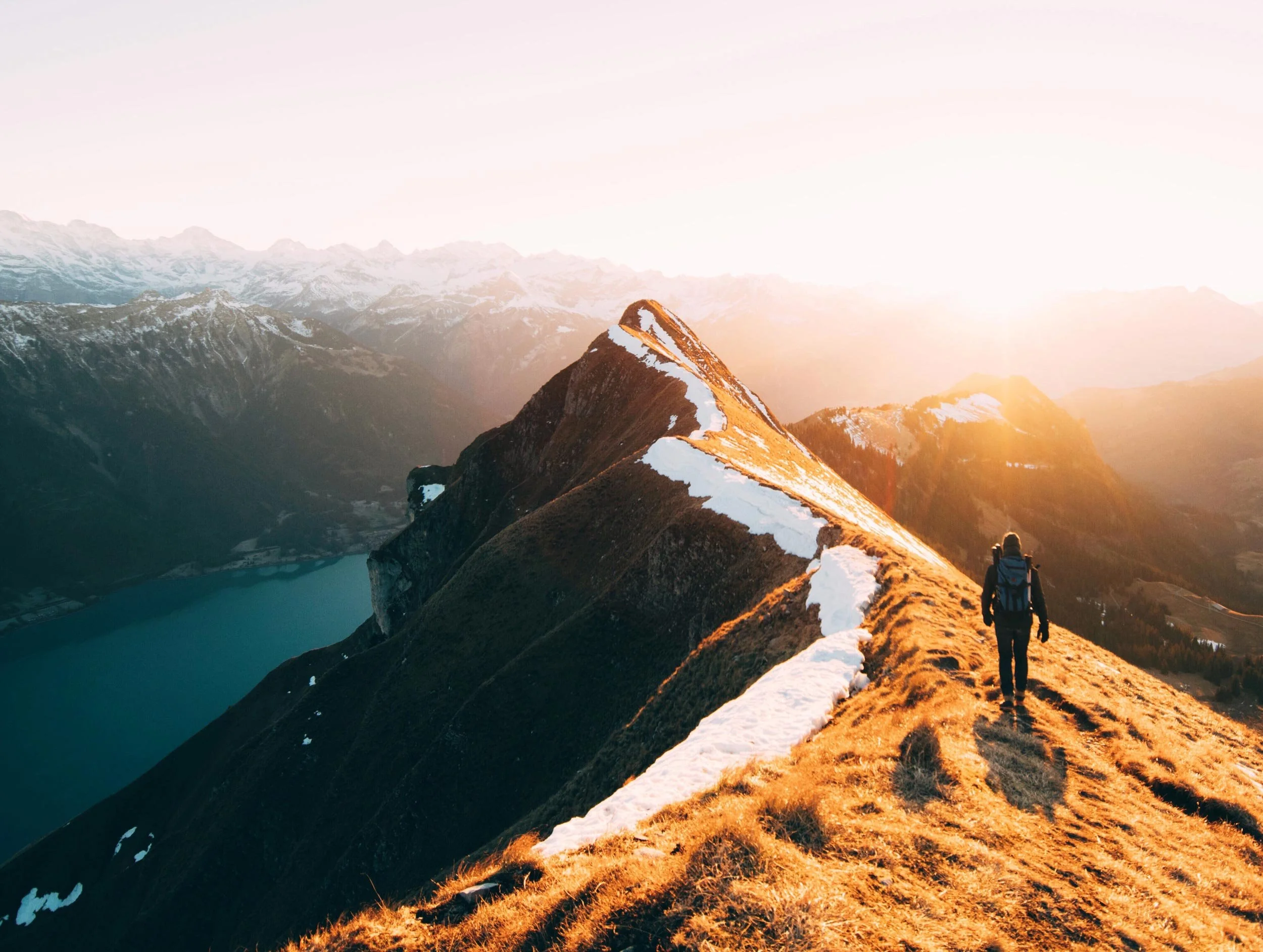 Solo hiker walking along a mountain ridge at golden-hour sunset with snowy peaks and a lake below