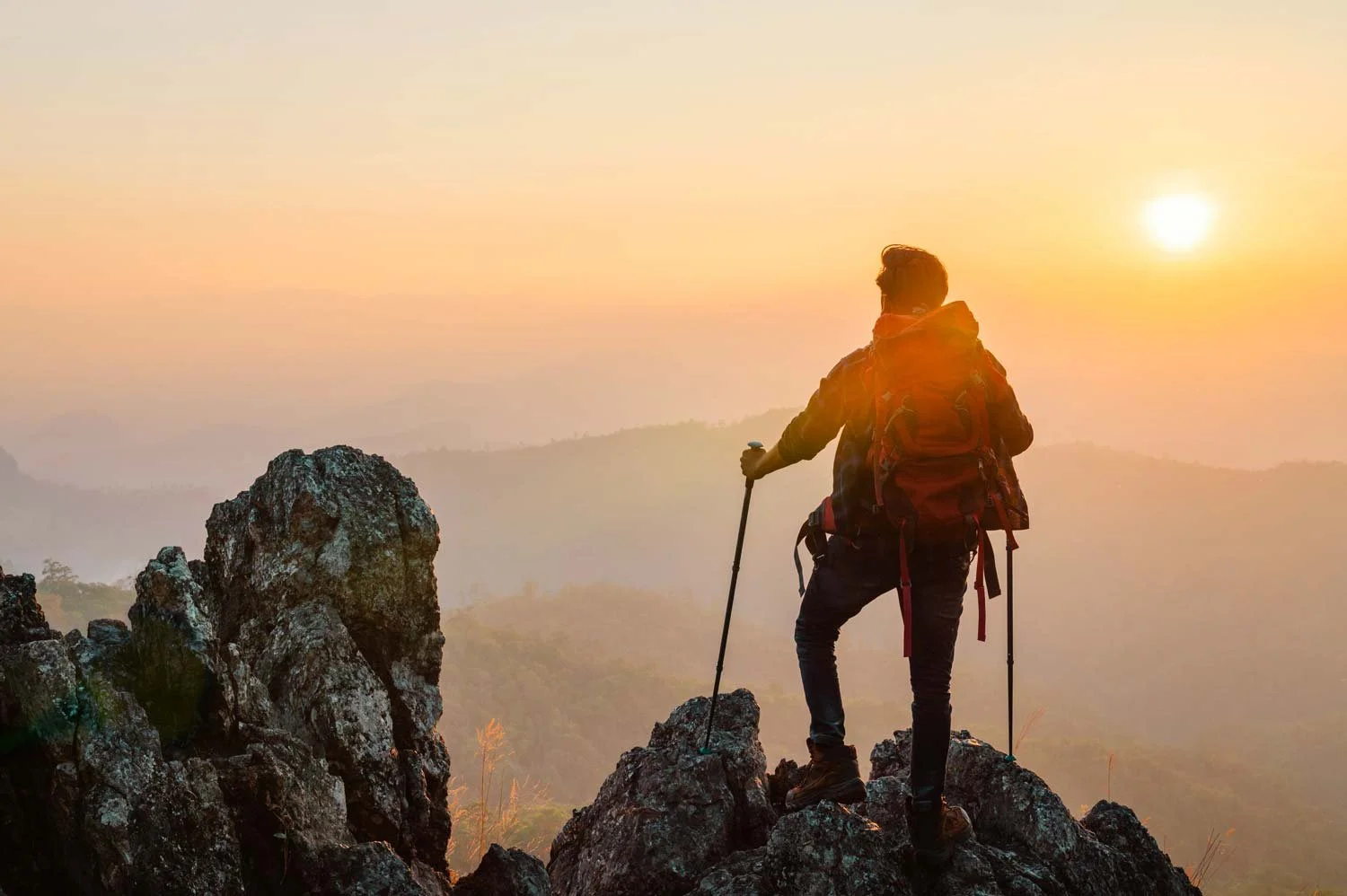 Backpacker on a rocky peak at warm sunset, backlit silhouette with trekking poles