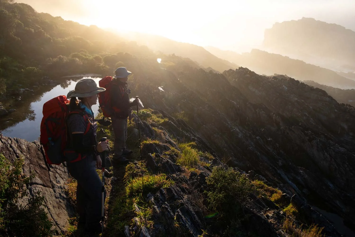 Two hikers with red backpacks standing on a rocky ridge in misty golden light