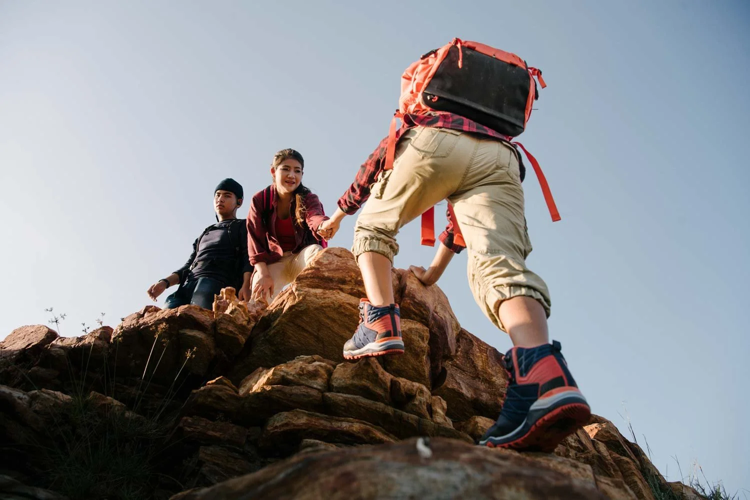 Hikers helping each other climb rocky boulders against a blue sky