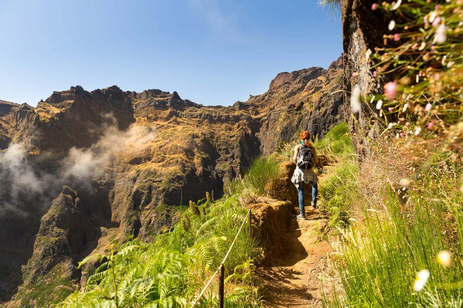 Female hiker on a narrow cliff trail surrounded by lush greenery and dramatic mountain walls