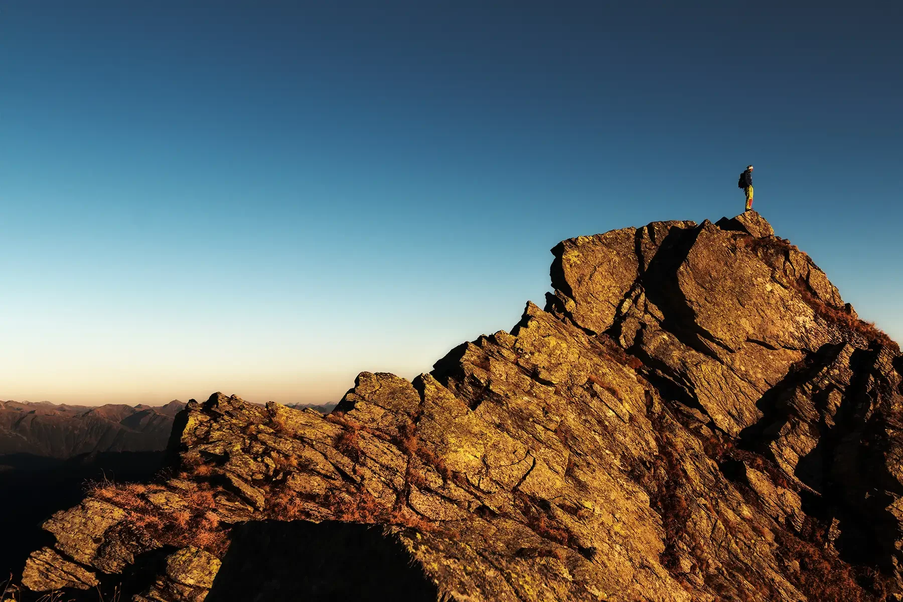 Lone hiker standing on a rocky mountain summit at sunset under a dramatic blue-orange sky
