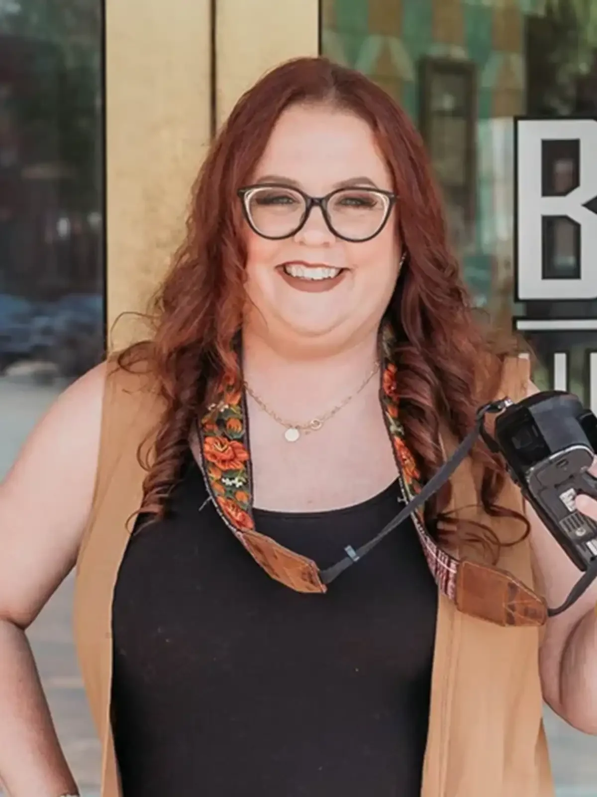 Amy Grissom, smiling with red curly hair and glasses, holding a camera outdoors