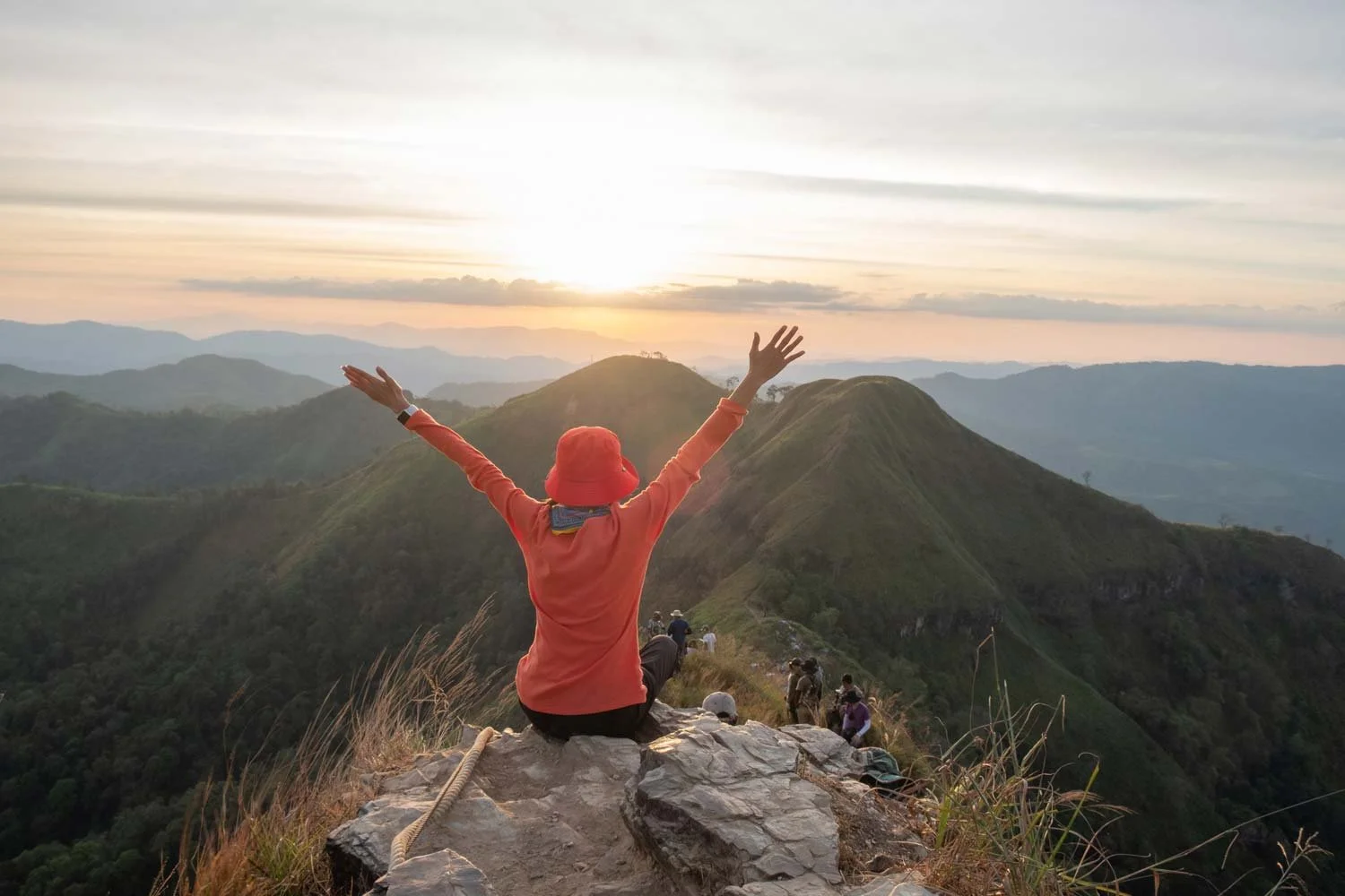 Person in an orange jacket with arms raised on a mountain summit at sunset