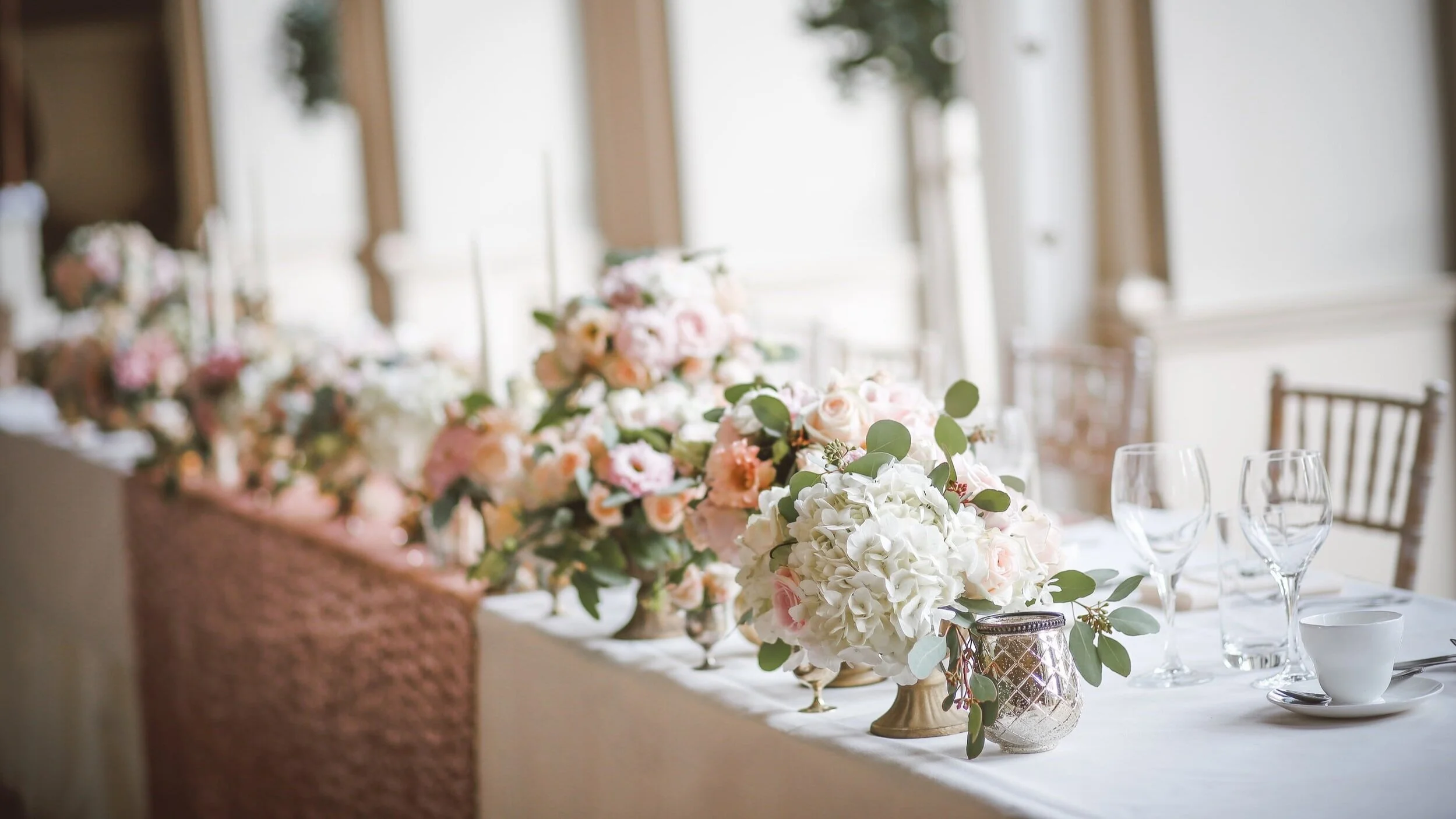 Elegant wedding reception head table with pink and white floral centerpieces, a champagne runner, and Chiavari chairs