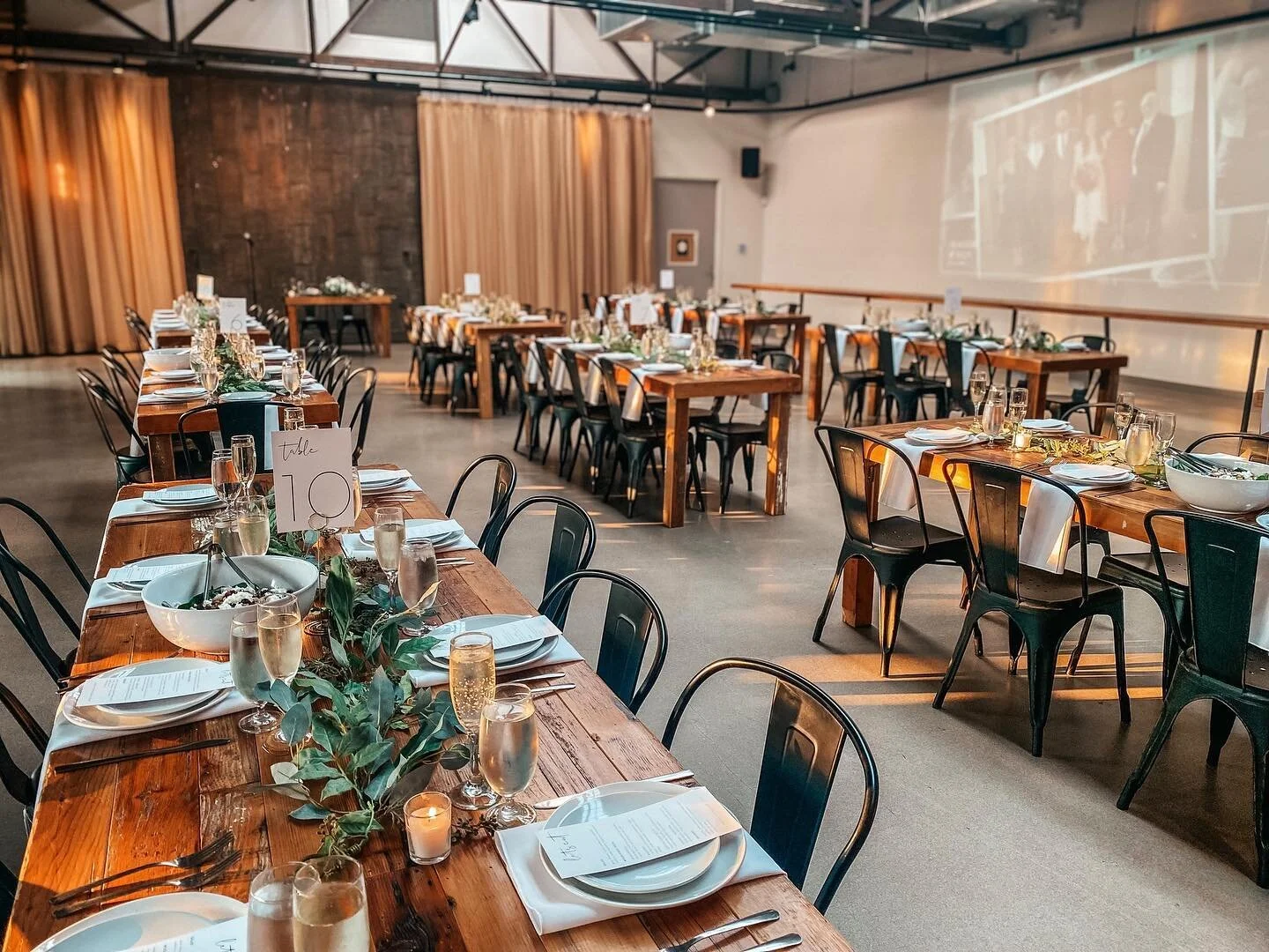 Industrial-style wedding reception hall with long farm tables and greenery runners