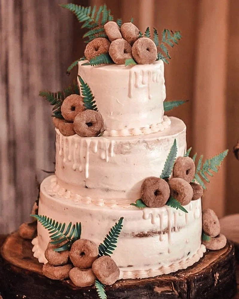 Three-tier naked wedding cake decorated with cinnamon sugar donuts and fern fronds on a wood stump stand