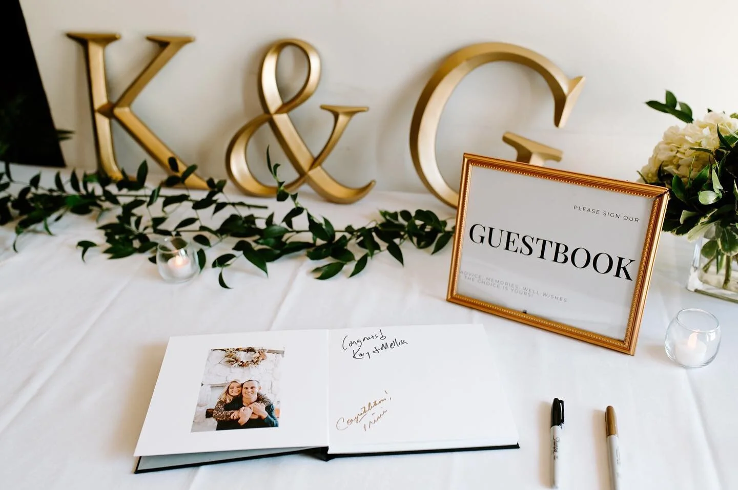 Wedding guestbook table with gold letter signs, a framed sign, and greenery