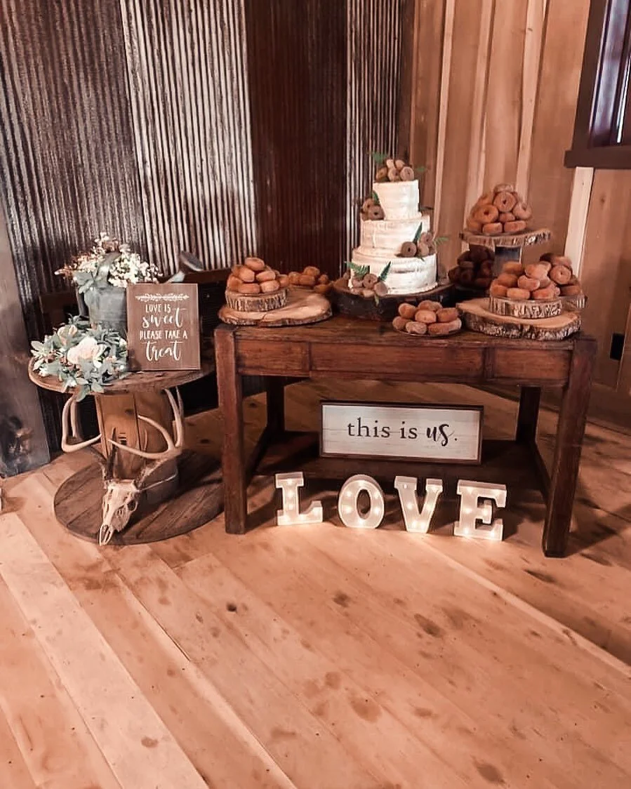 Rustic barn wedding dessert table with a tiered naked cake, donuts on wood slices, LOVE marquee letters, and a 'this is us' sign