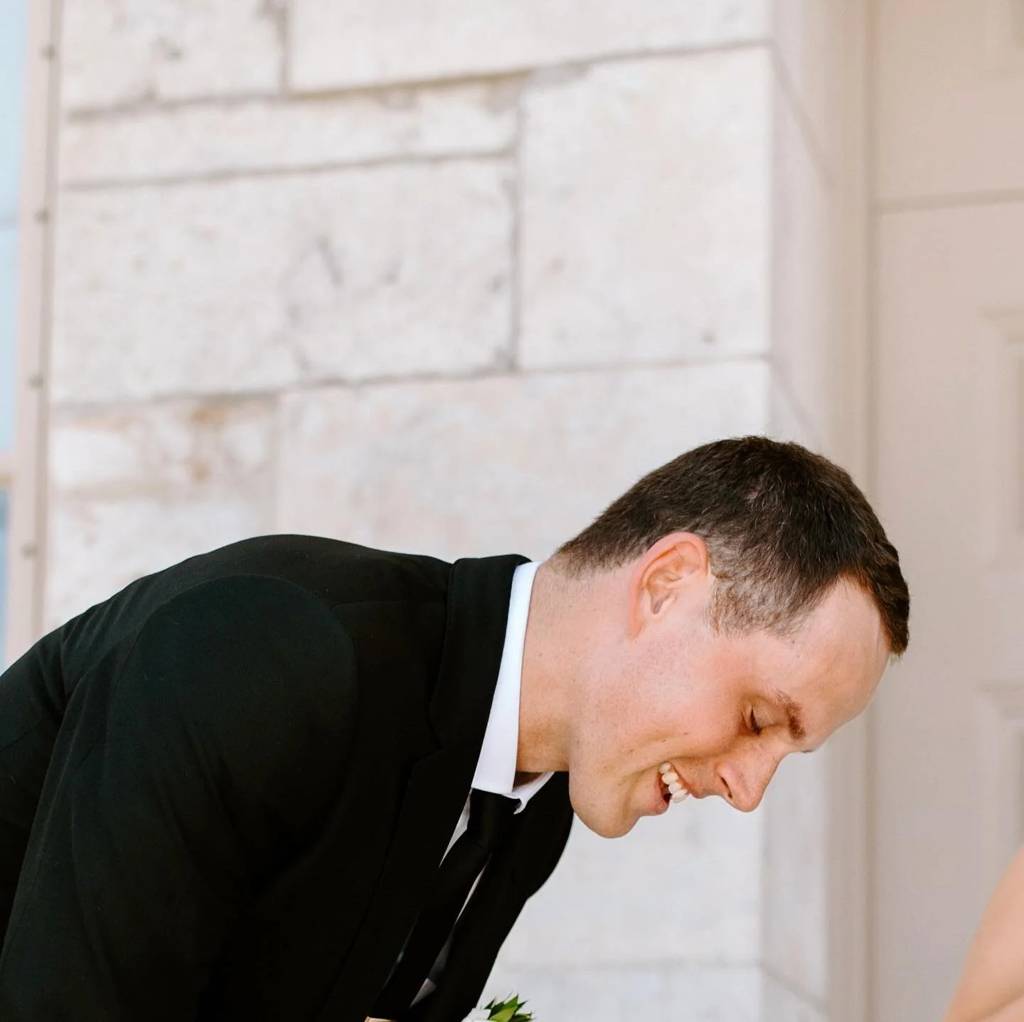 Groom in a black tuxedo laughing in front of a stone-wall venue