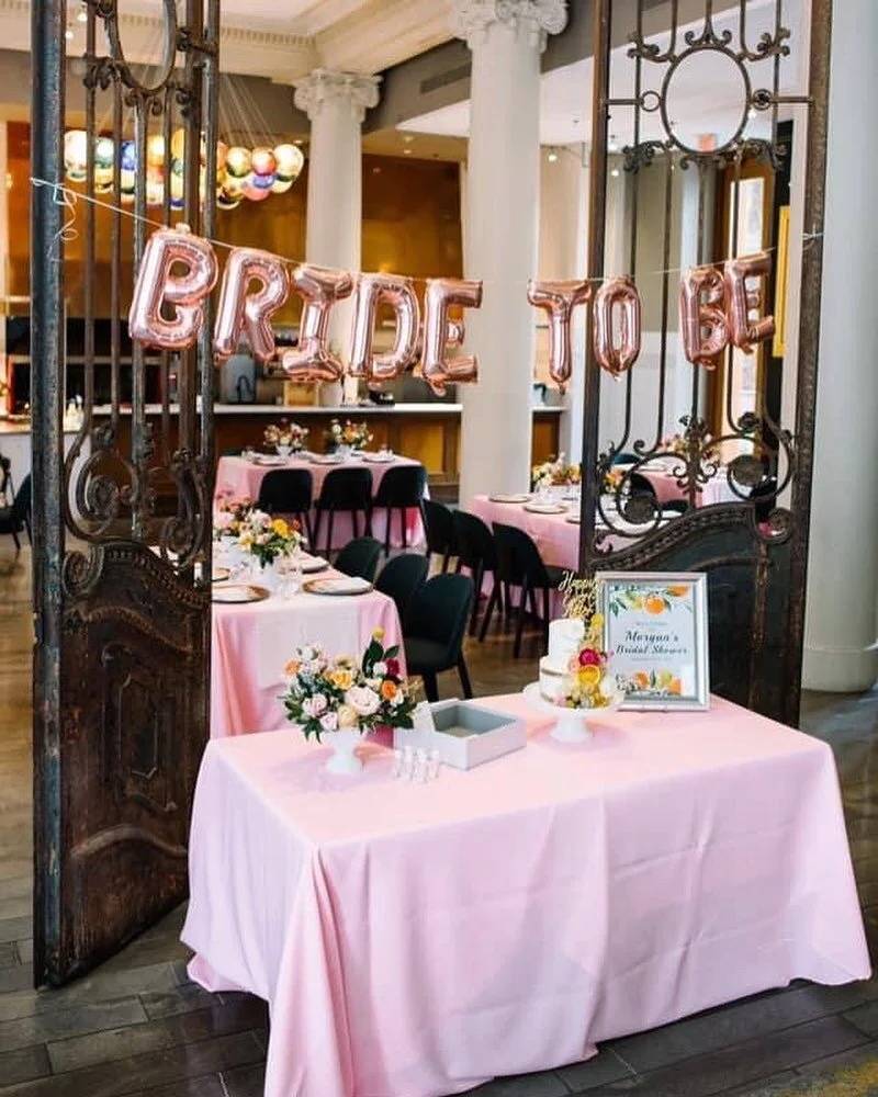 Bridal shower setup seen through ornate iron gates with 'BRIDE TO BE' balloon letters