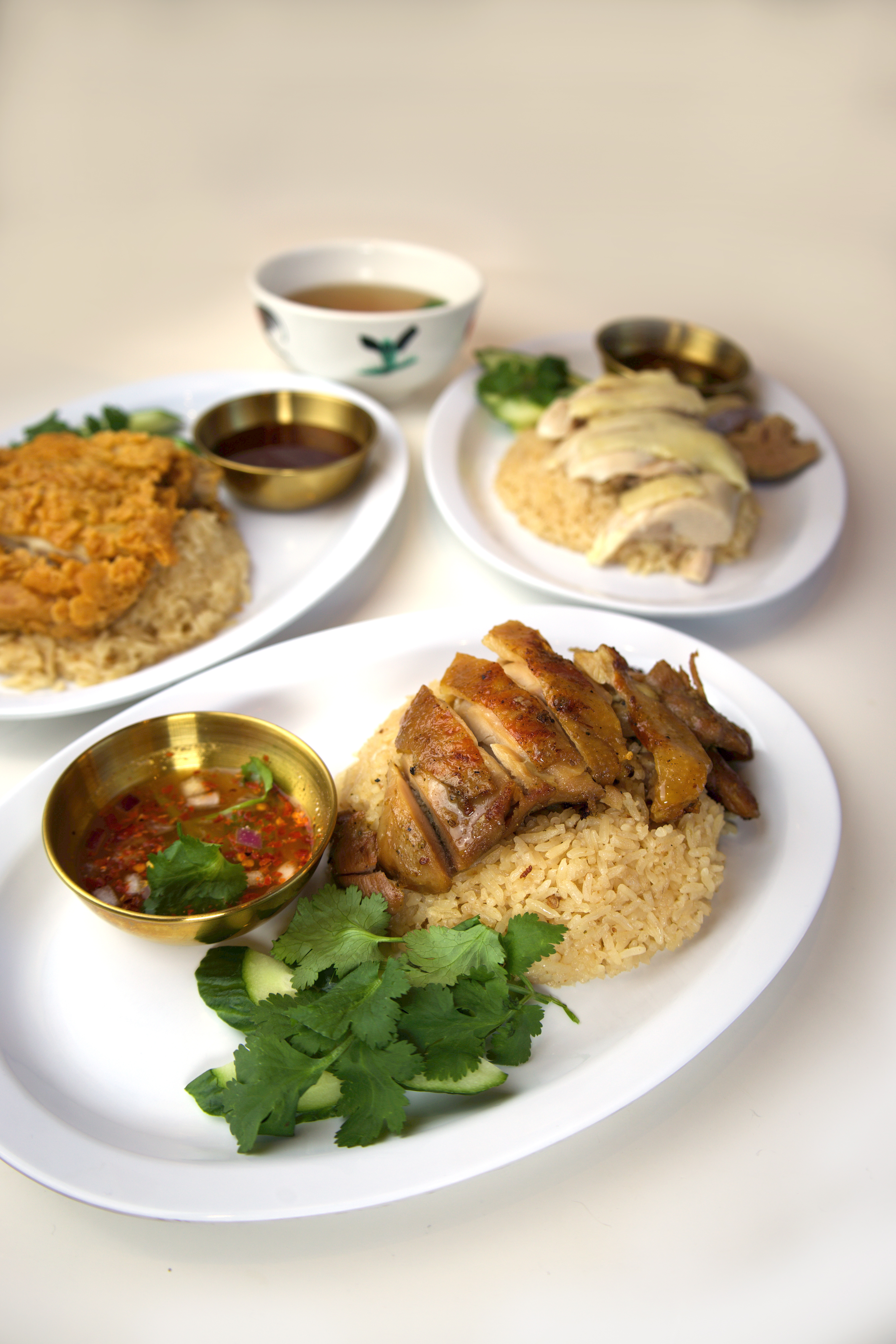 Three plates of Thai chicken rice — roasted, poached and fried — with gold bowls of dipping sauce