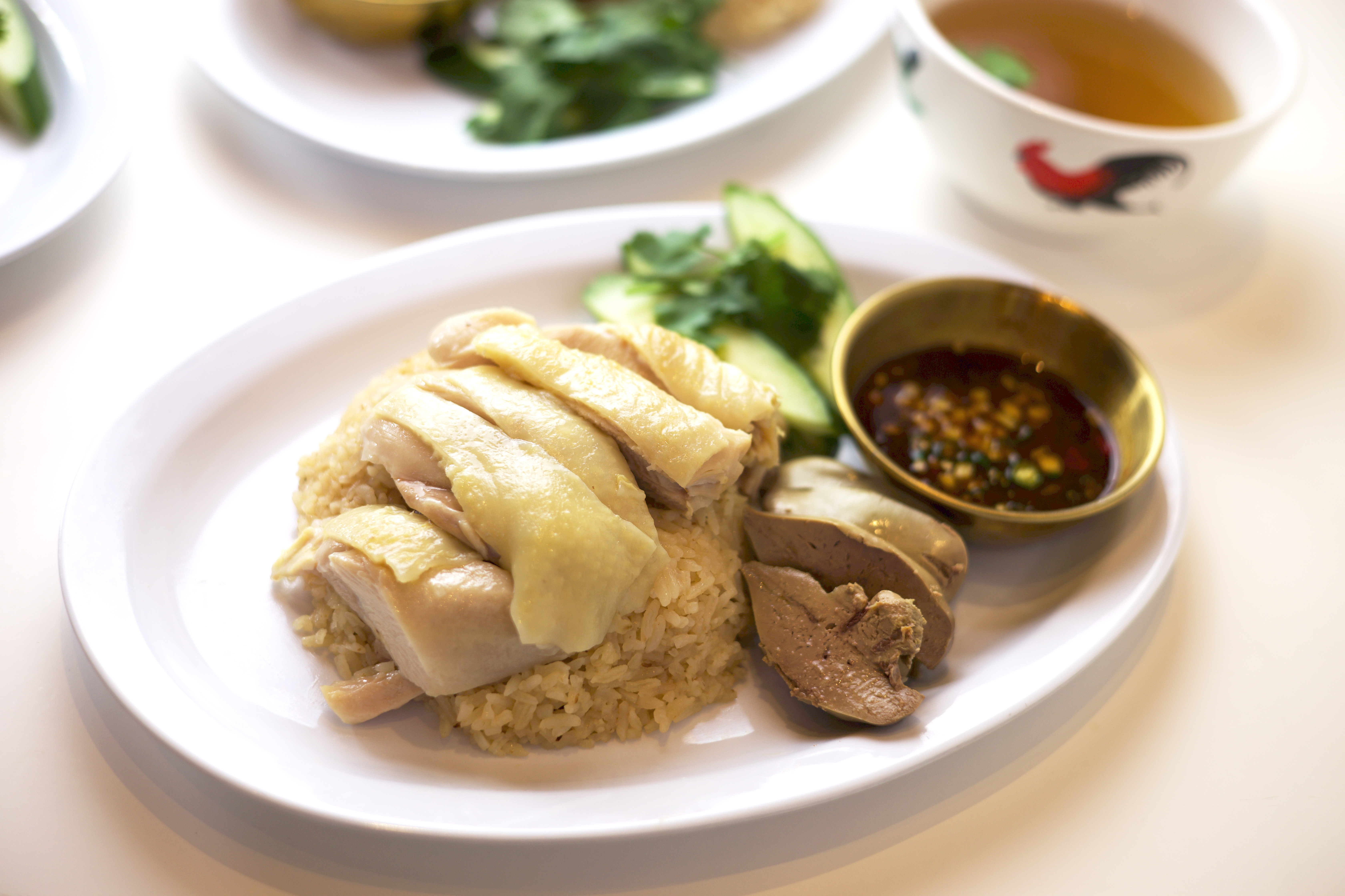 Close-up of poached Khao Man Gai with sliced chicken, dark dipping sauce, cucumber and a cup of broth