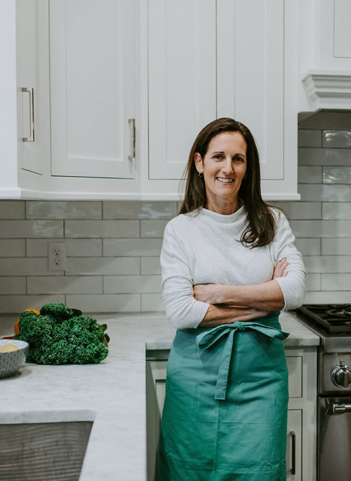 Heather Carey standing with arms crossed in a white kitchen, wearing a teal apron