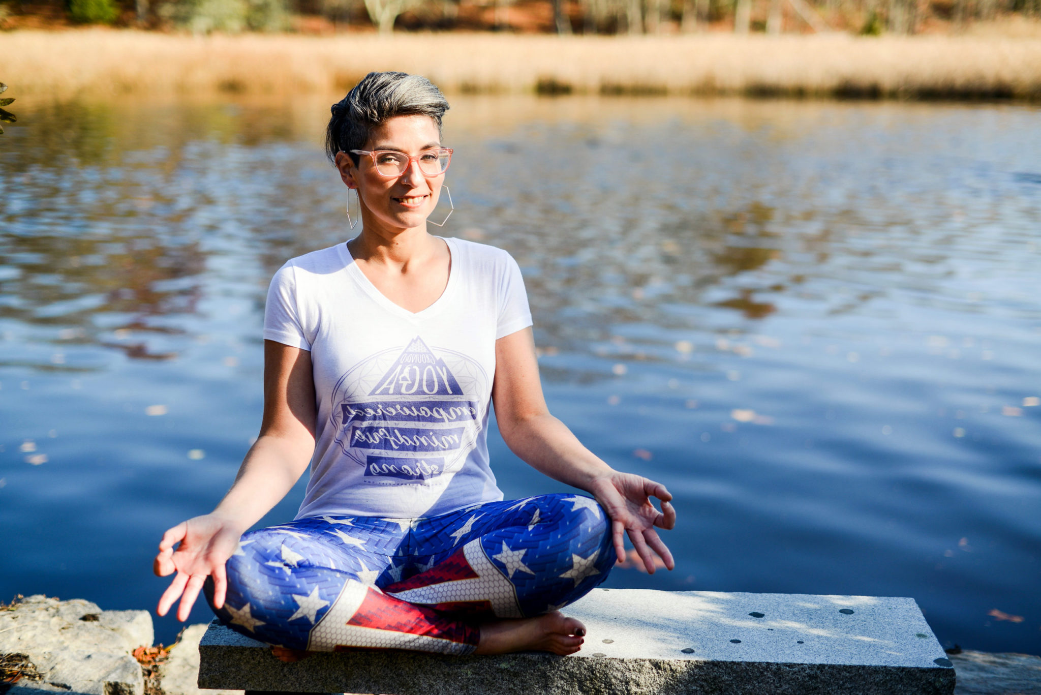 Rachel Dash-Dougherty meditating in lotus pose on a stone ledge beside a Connecticut lake.