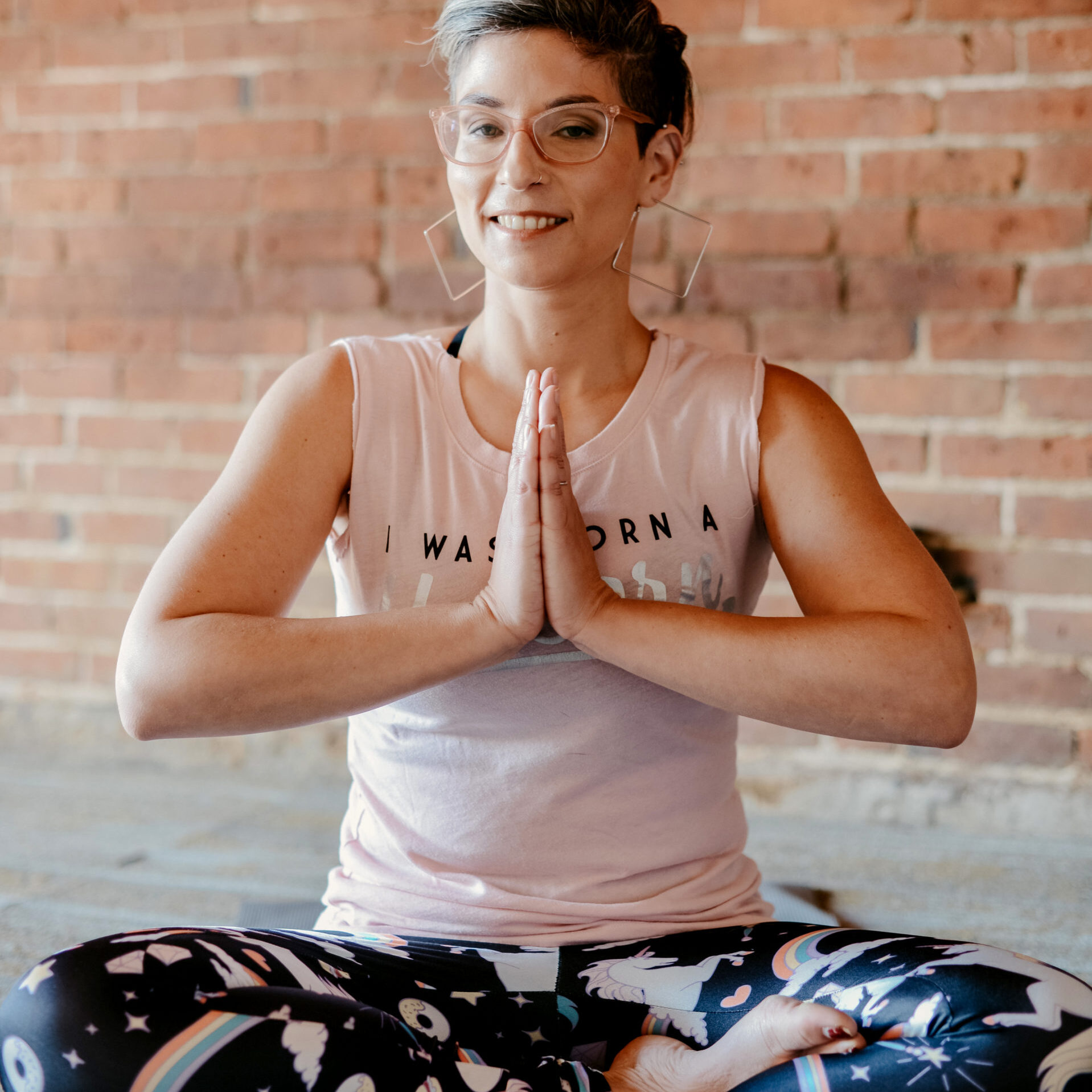 Rachel Dash-Dougherty seated cross-legged in yoga prayer pose against a brick wall.