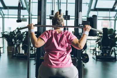 A woman seen from behind in a pink t-shirt performing a lat pull-down on a gym machine