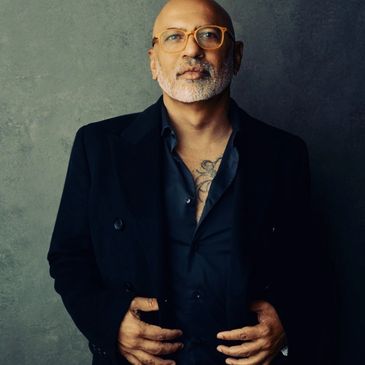 Studio portrait of Vineet Thapar, a bald man with a beard and amber-framed glasses wearing a dark blazer