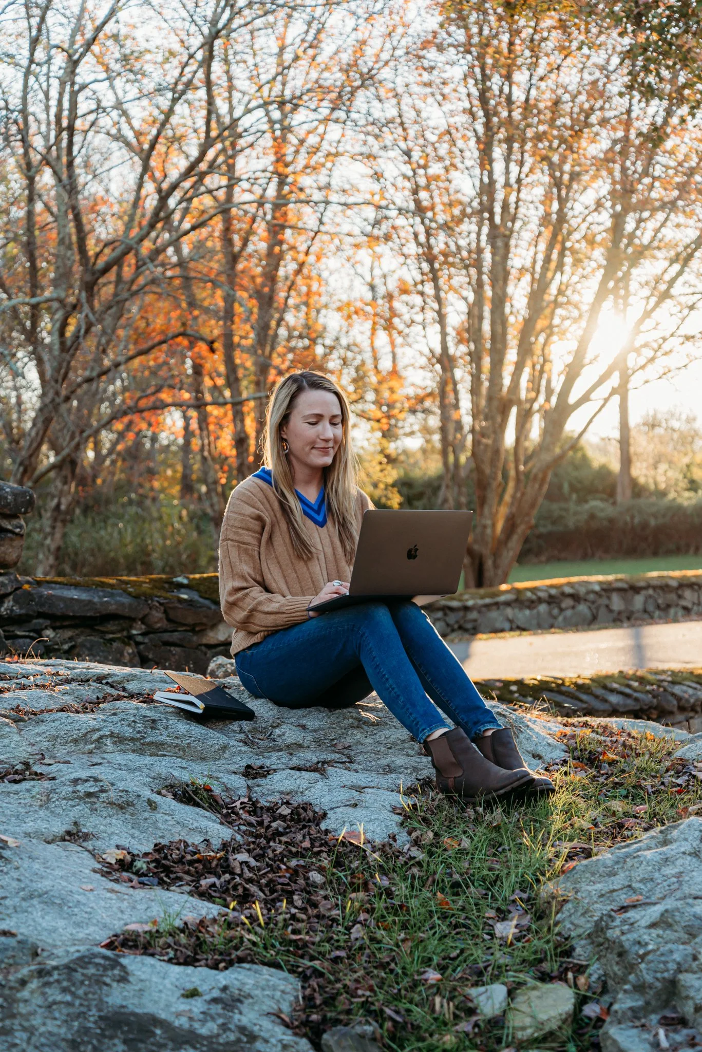 Hannah seated on a rock outdoors with a MacBook, autumn foliage in the background.