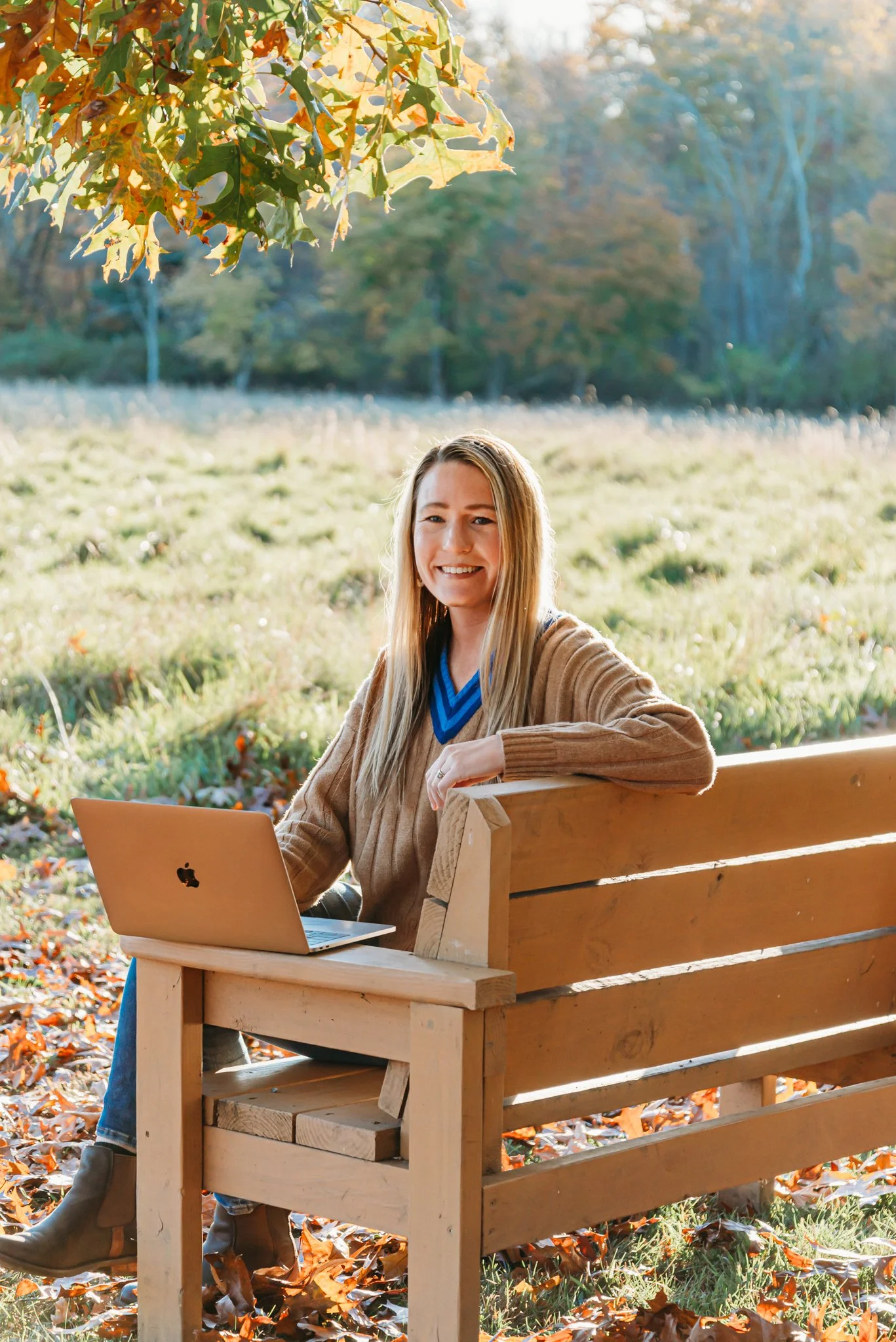Hannah Morton smiling, seated at a wooden bench with a MacBook in an autumn field.