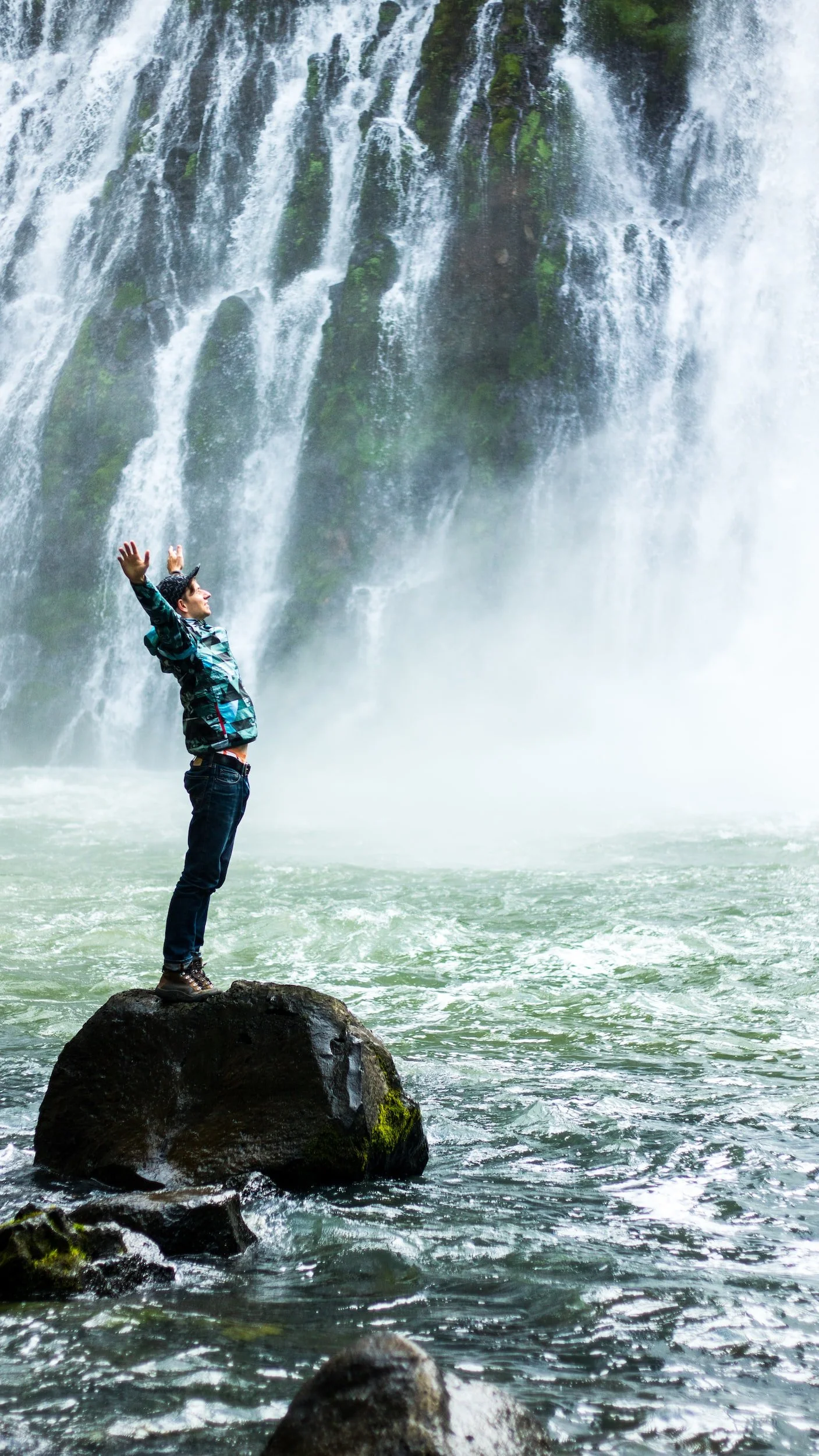 Person standing on a rock with arms raised in front of a large waterfall.