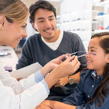 Optician fitting eyeglasses on a young girl while her father looks on