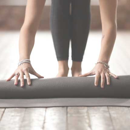 Close-up of a woman's hands and feet rolling up a grey yoga mat on a wooden floor
