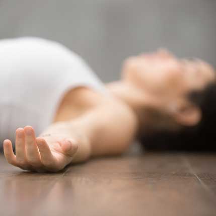Woman lying in savasana on a wooden floor with a relaxed open hand in the foreground