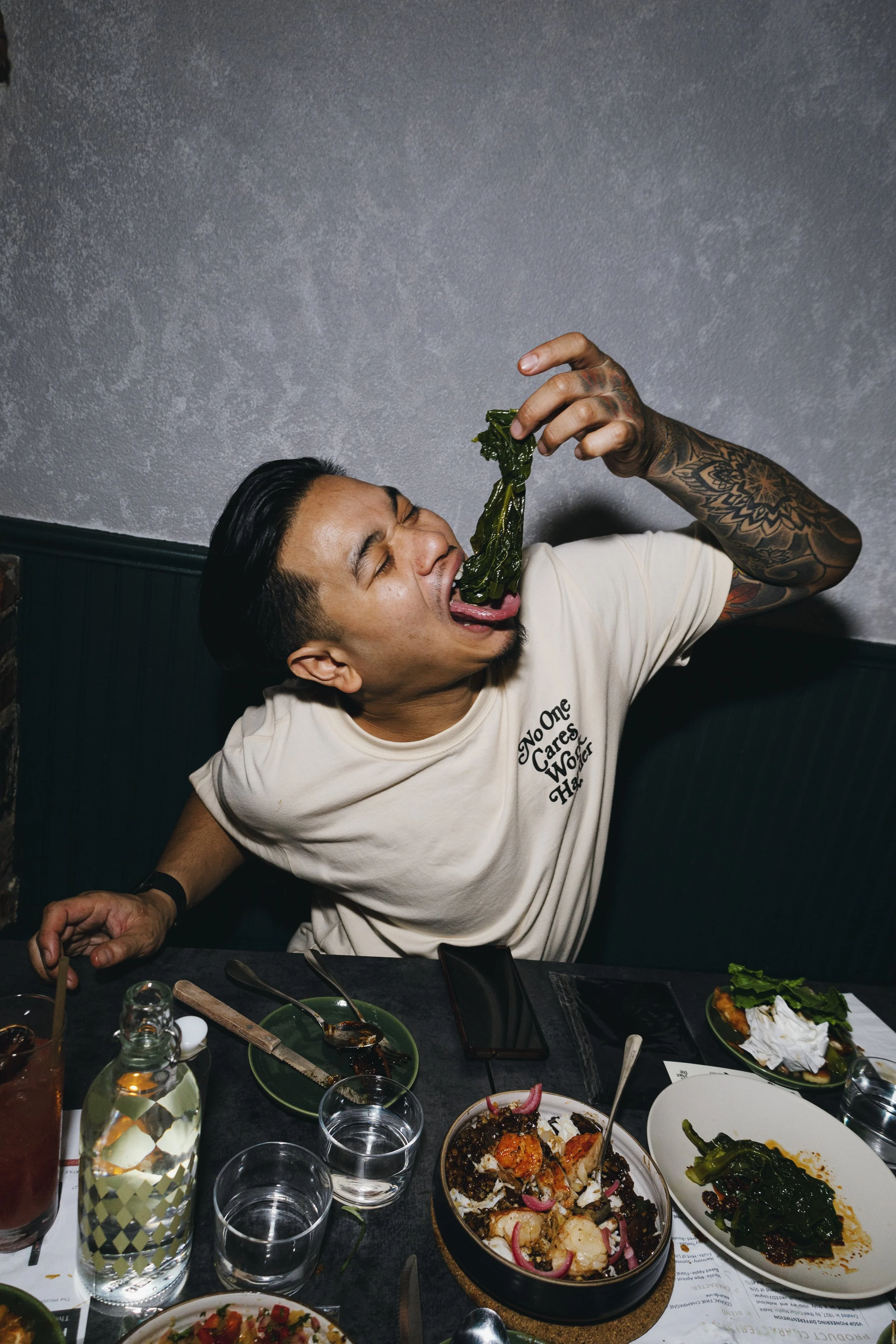 A chef at a restaurant table, eating greens by hand surrounded by shared dishes.