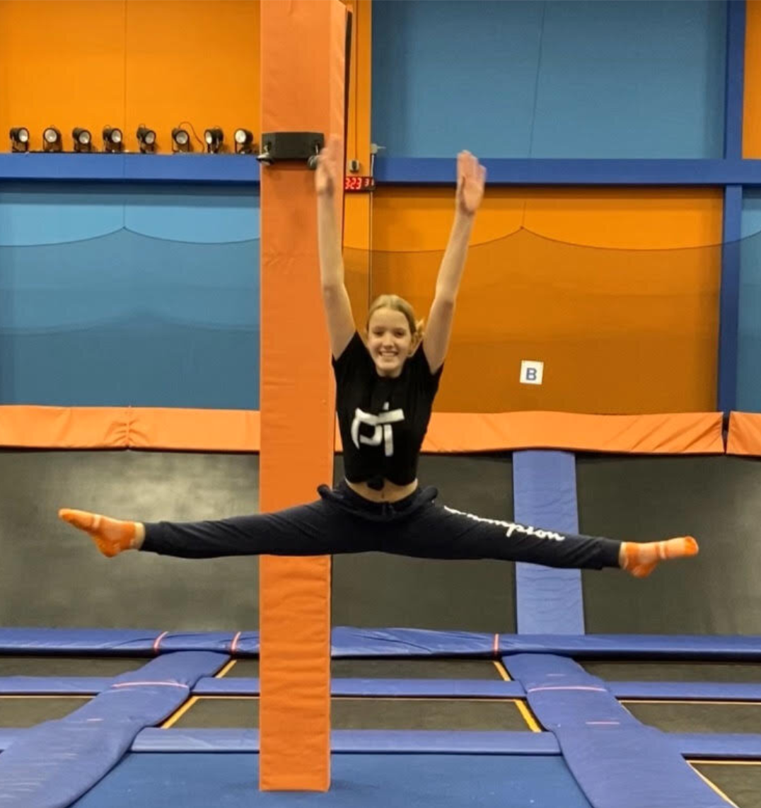 Kate R in a black House of Physical Therapy shirt doing a mid-air split jump at an indoor trampoline park