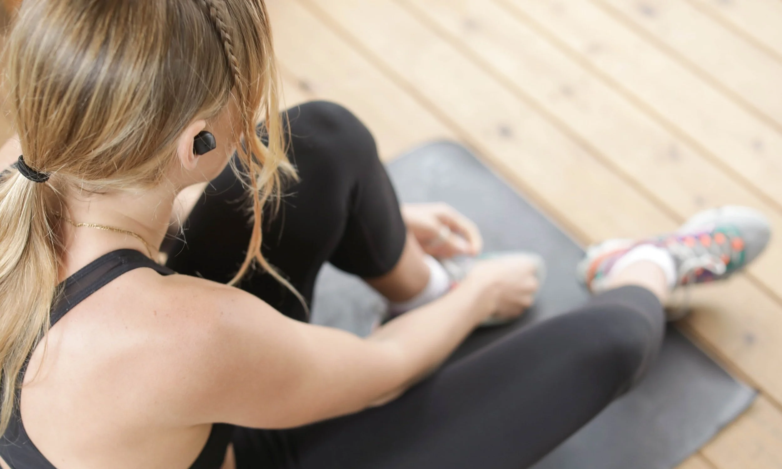 Overhead view of a woman in athletic wear sitting on a gym mat, adjusting her shoe on a wood floor