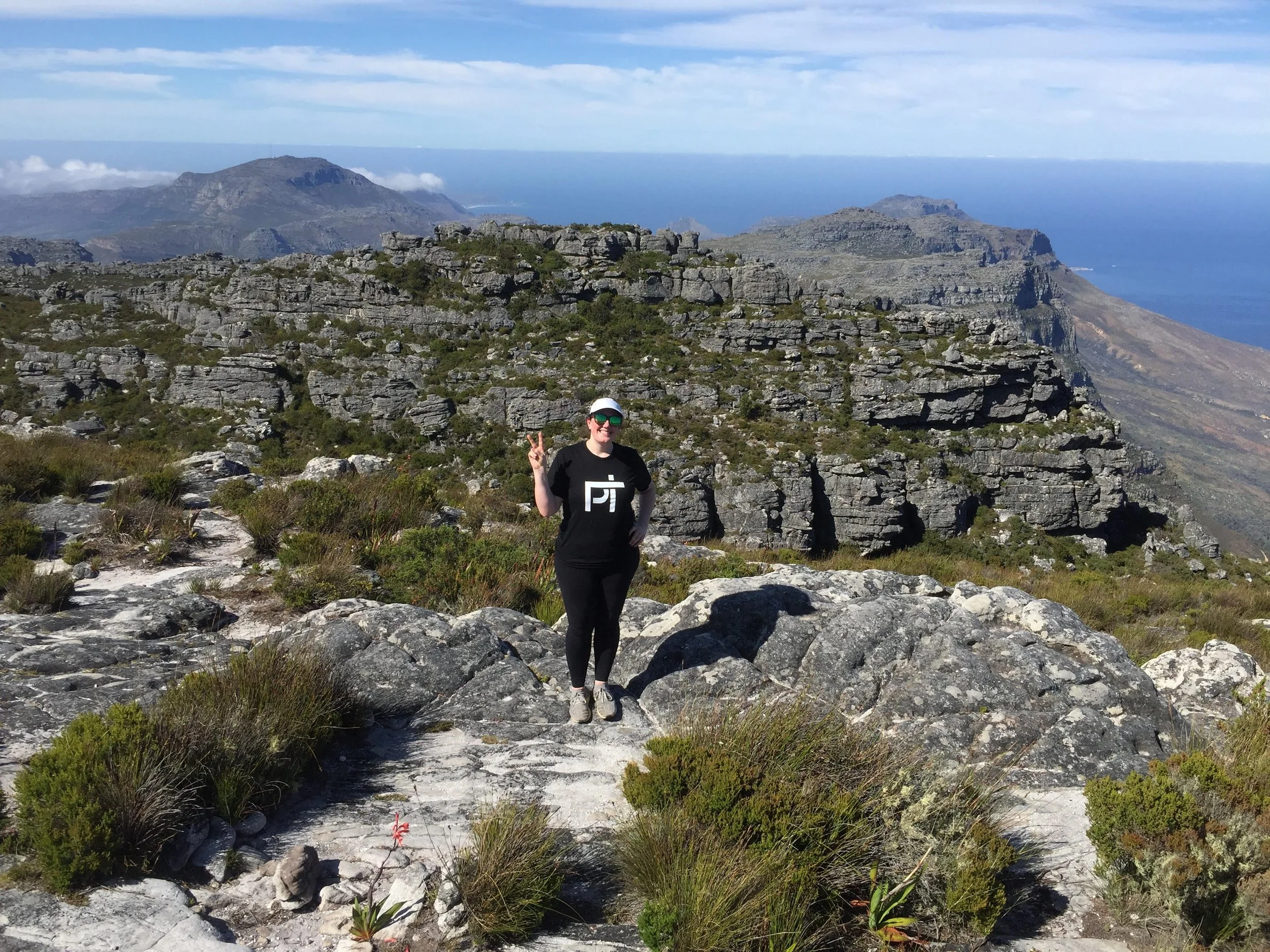 Beth in a House of Physical Therapy shirt on a rocky mountain summit above the ocean in South Africa