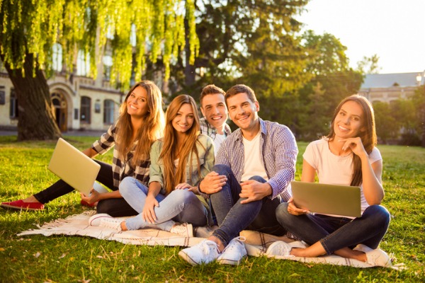Five smiling young adults sitting on grass with laptops