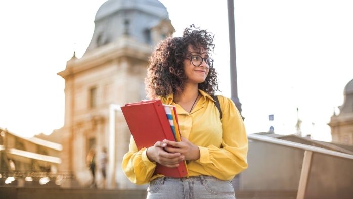 Young woman with curly hair and glasses holding books outdoors near a historic building