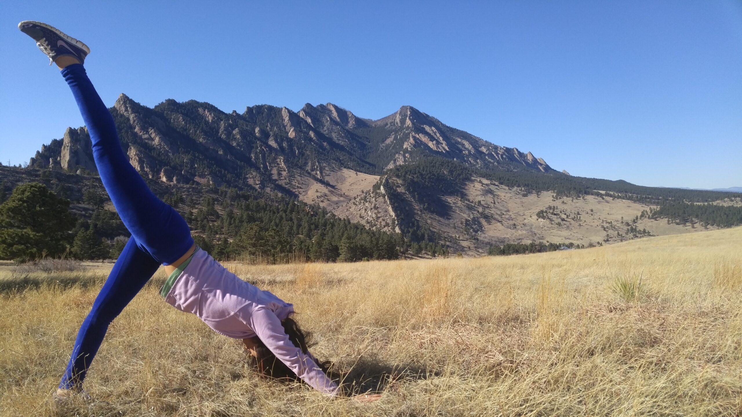 Penpa Dolma in a one-legged downward dog pose in a Colorado meadow with the Flatirons behind