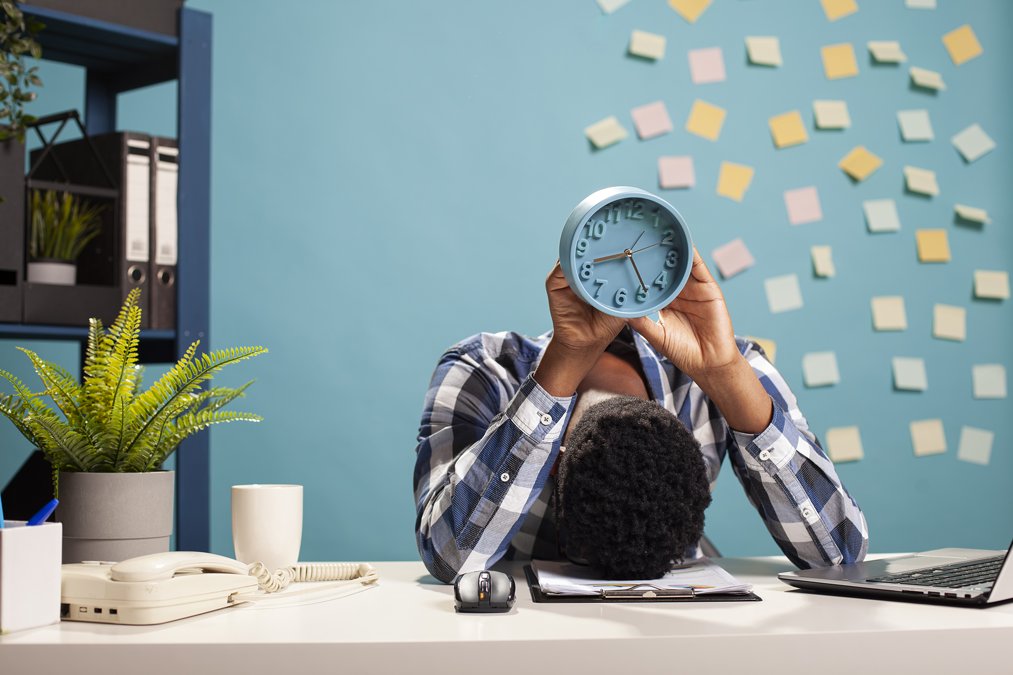 A person slumped over a desk holding a clock — a burnout concept
