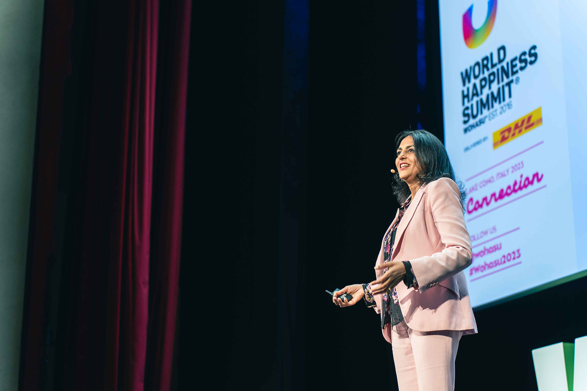 Dr. Neha Sangwan in a pink blazer speaking on stage at the World Happiness Summit, Lake Como 2023