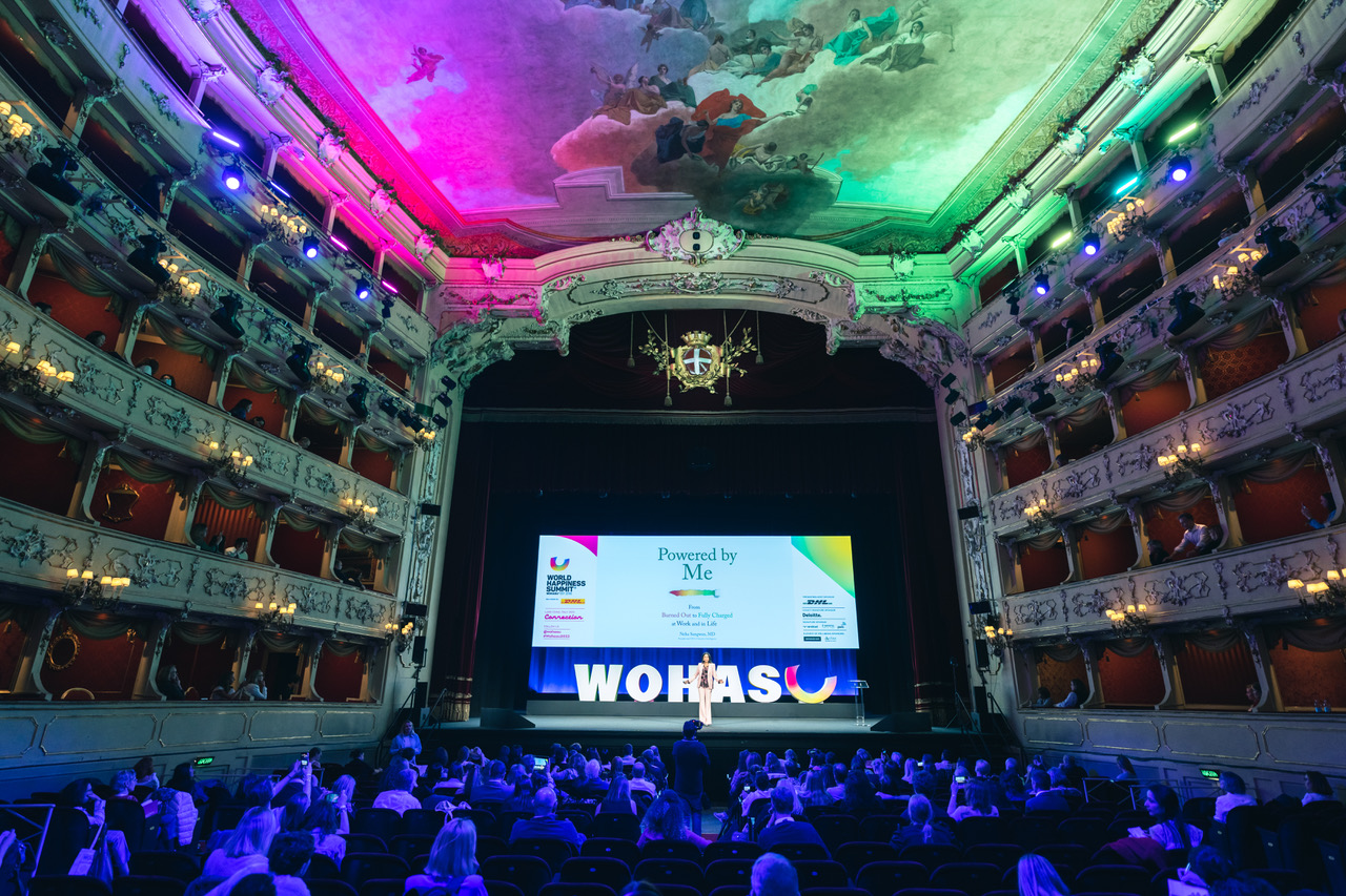 Ornate Italian theater with audience and Powered by Me on the stage screen at the World Happiness Summit