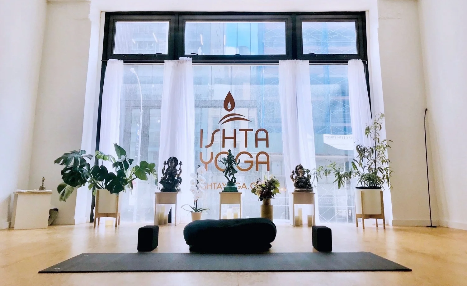 Bright ISHTA Yoga studio interior with the ISHTA logo on the window, a mat and bolster, Hindu statues and plants near Union Square in NYC