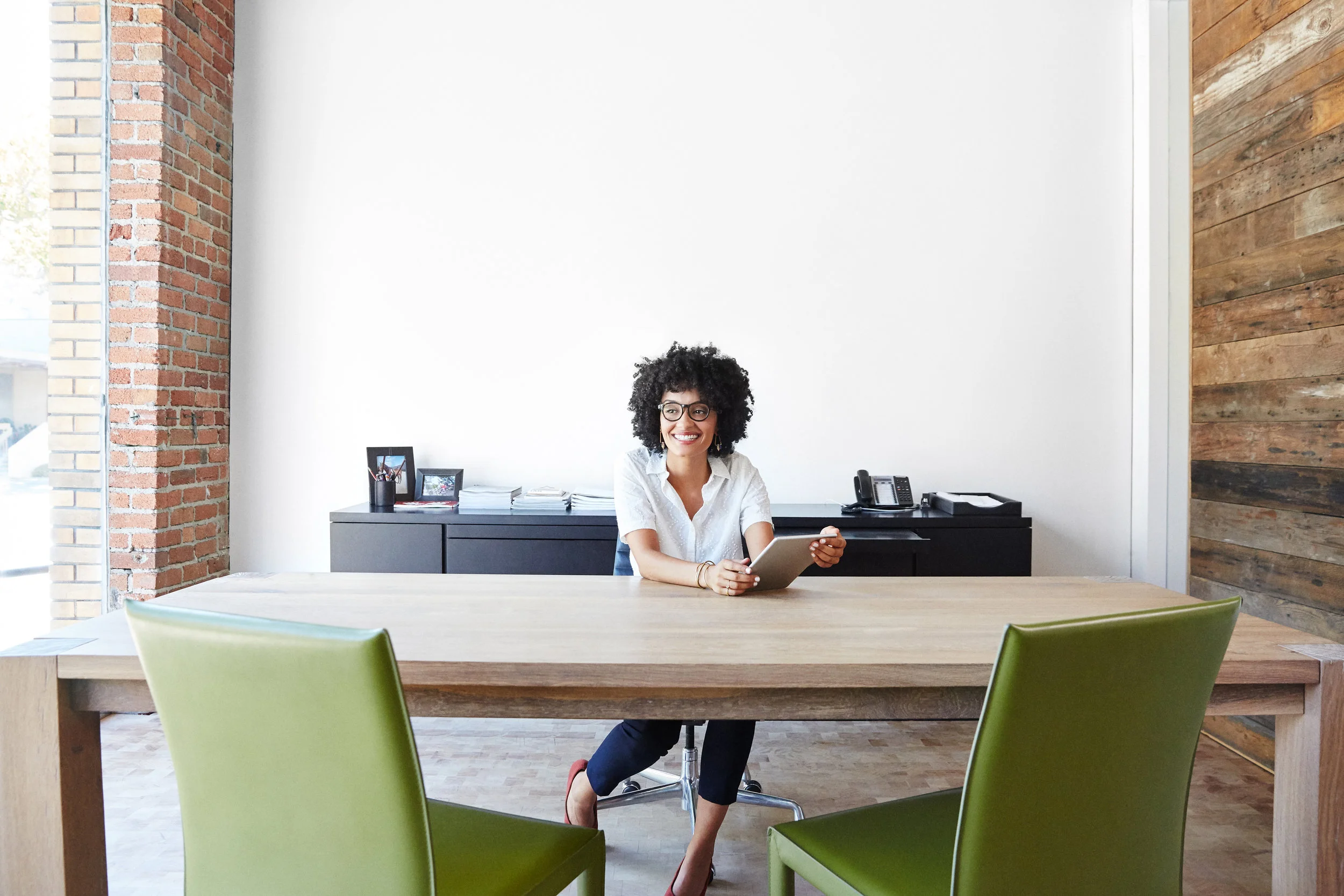 DEI consultant with natural curly hair and glasses sitting at a conference table in a modern office with brick and wood-panel walls, holding a tablet