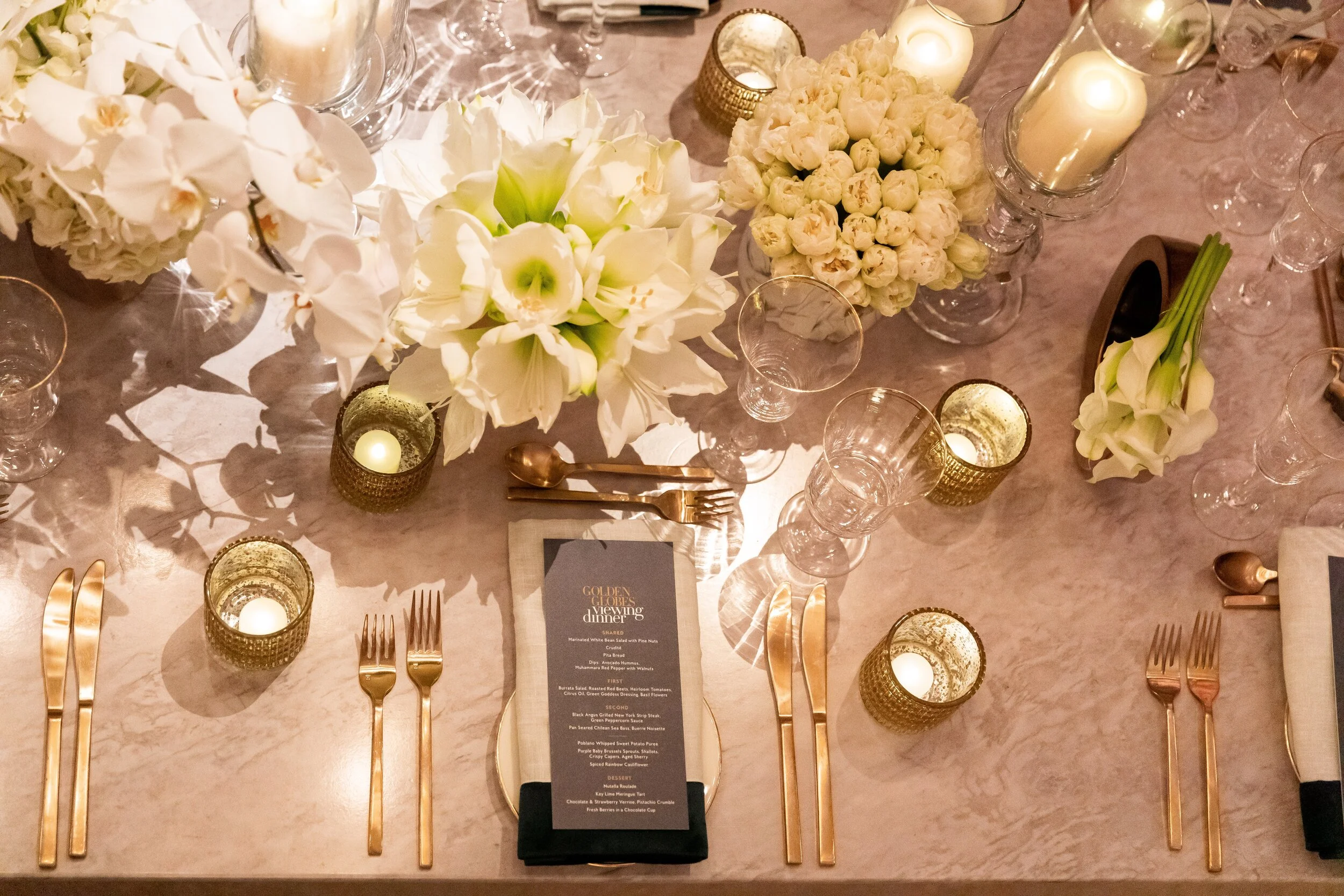 Overhead of an elegant Golden Globes Viewing Dinner table setting with gold cutlery, white flowers, candles and a printed menu card