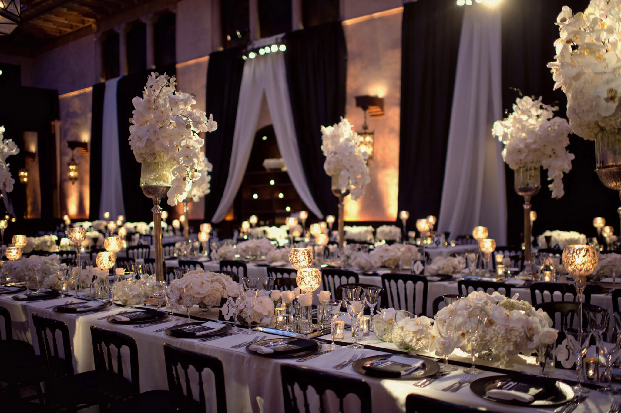 Grand ballroom set for a formal event with tall white orchid centerpieces, candlelight and dramatic black and white drapery