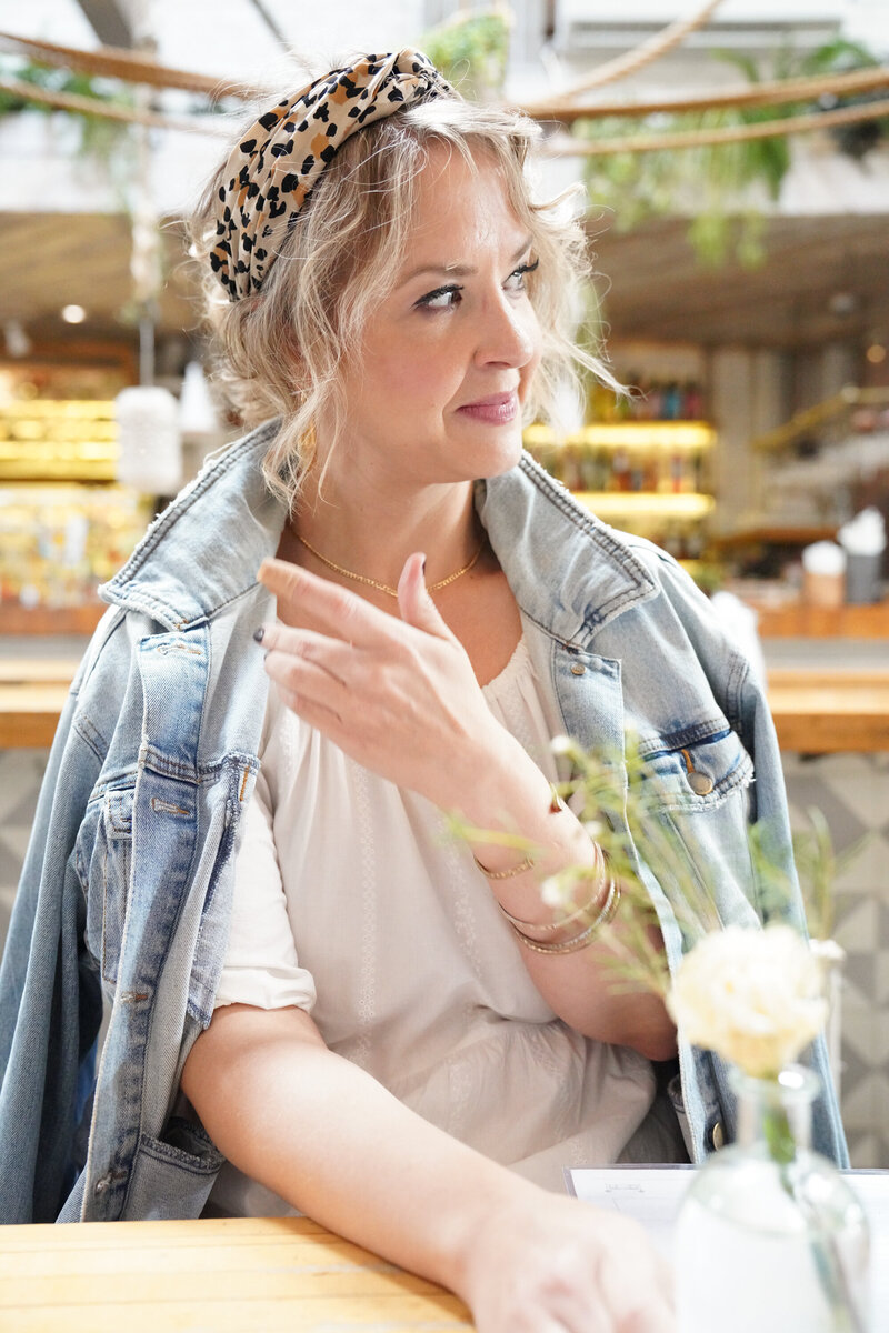 Jacqueline with a leopard headband seated at a cafe table, looking away thoughtfully
