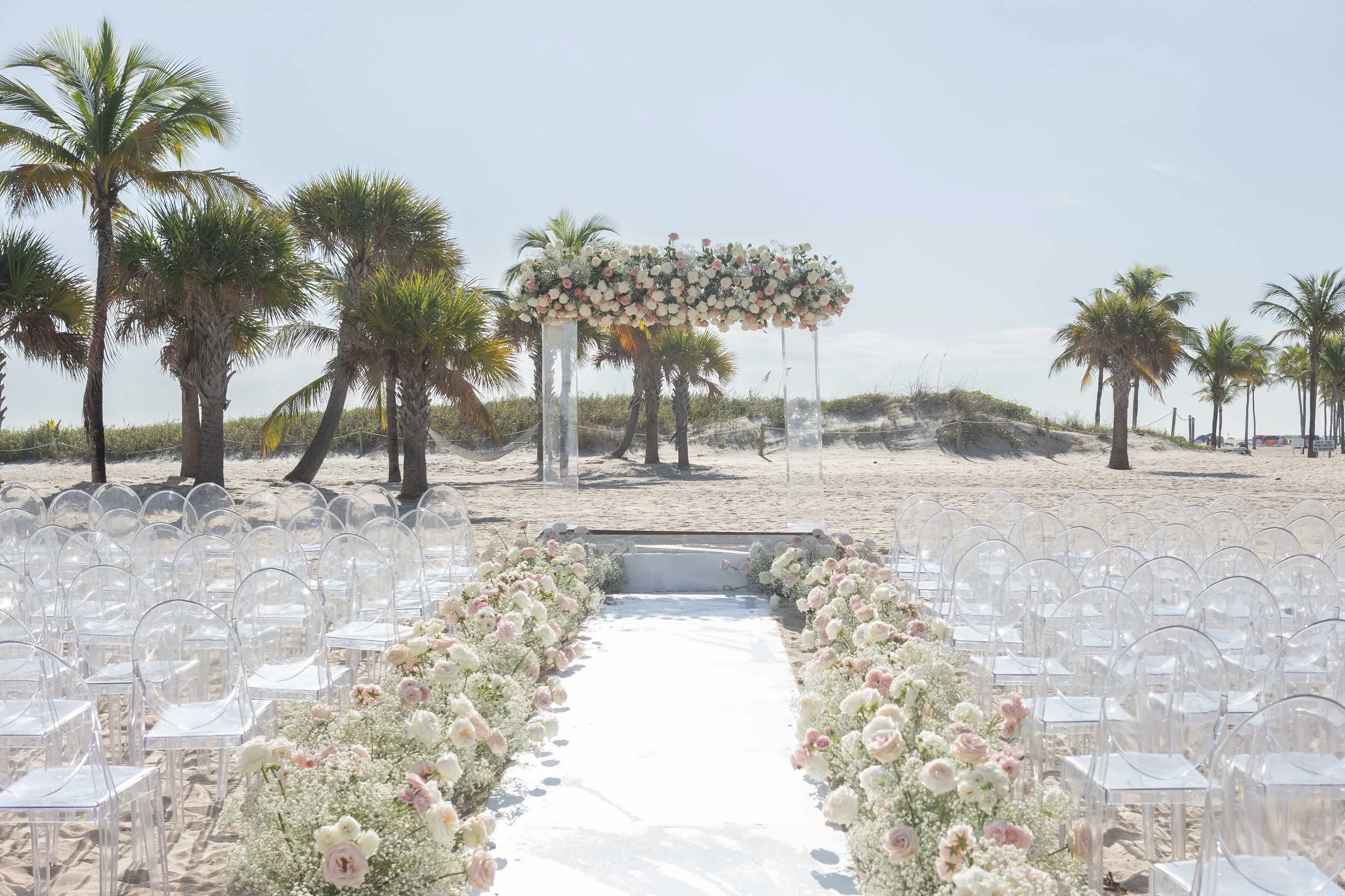 Beach wedding ceremony with a white aisle runner, ghost chairs and a floral arch under palms