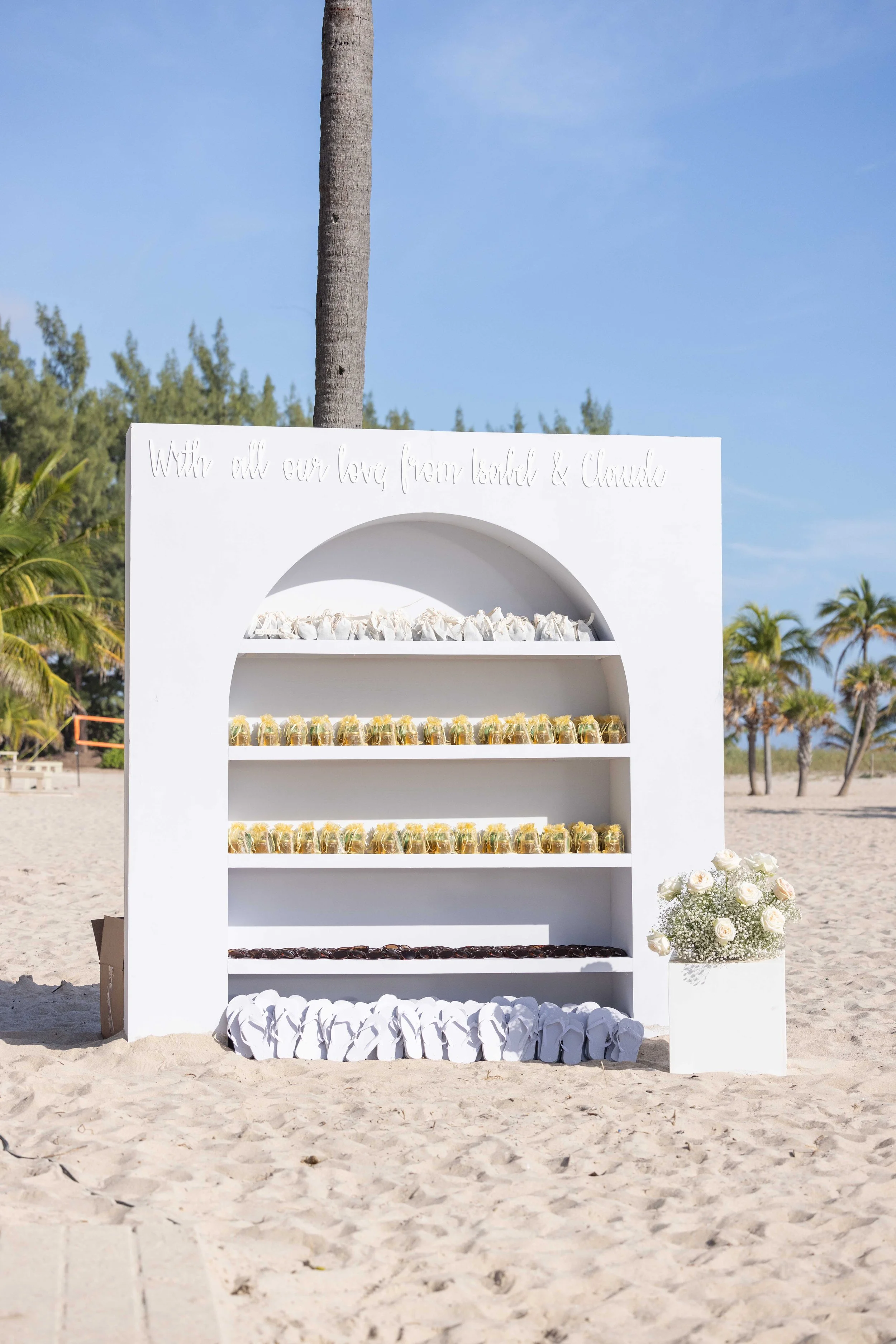 Custom white arch-shaped wedding favor display on sand with flip flops and gifts