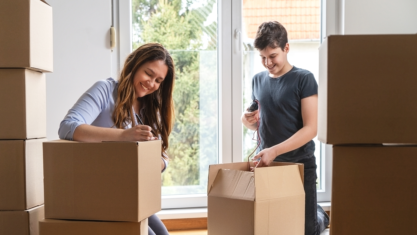 A woman and a teenage boy packing moving boxes in a bright, sunlit room