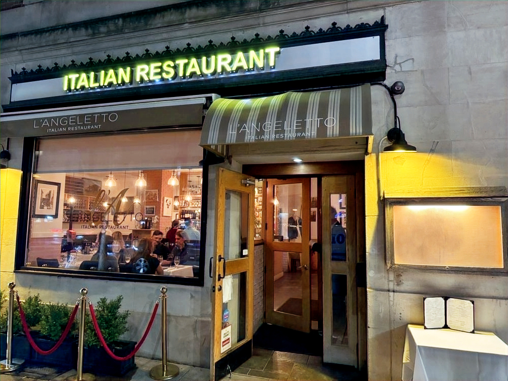 L'Angeletto Italian Restaurant storefront at night with illuminated signage, striped awning and diners visible in the window
