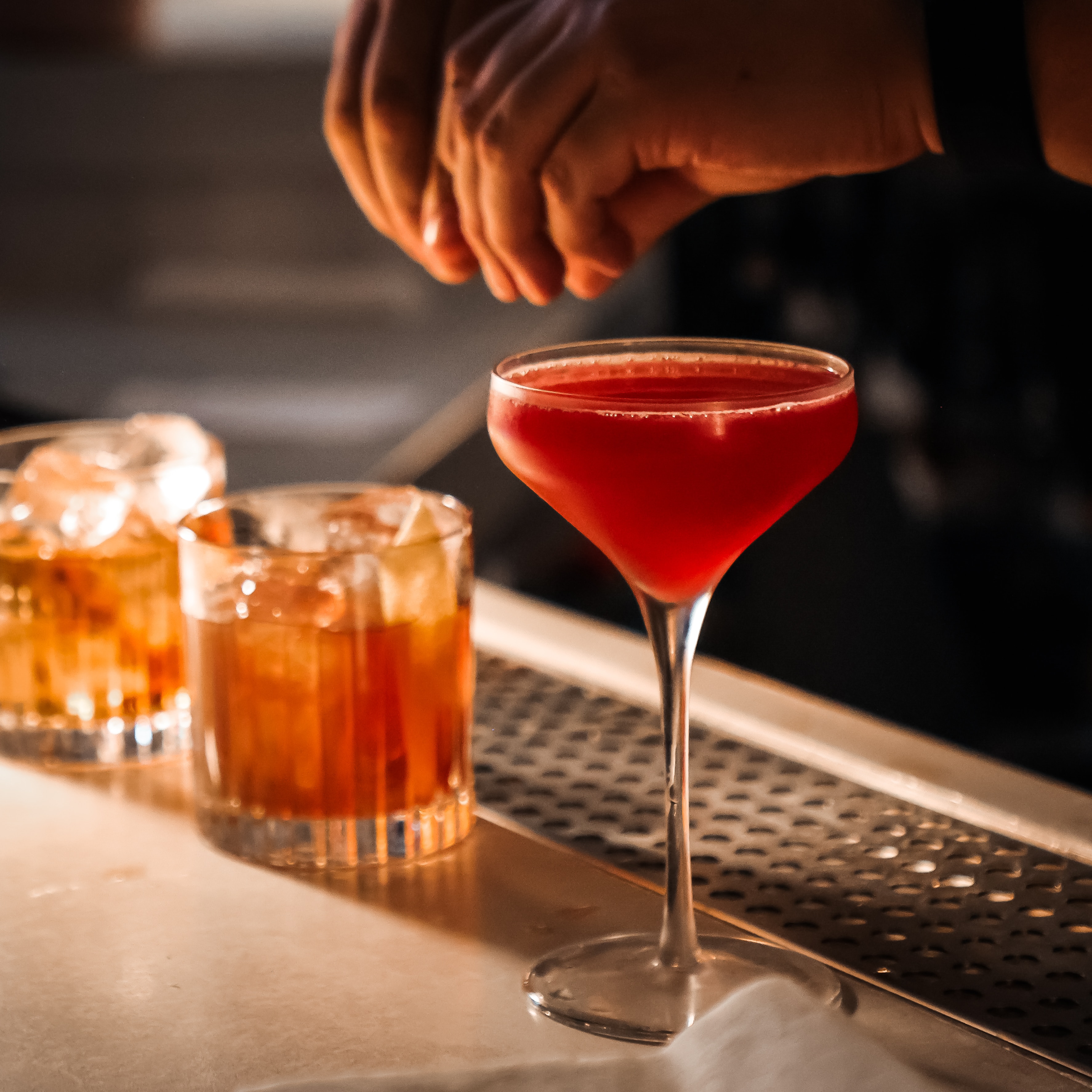 Bartender finishing a red cocktail in a coupe glass beside two amber whiskey glasses on the bar rail